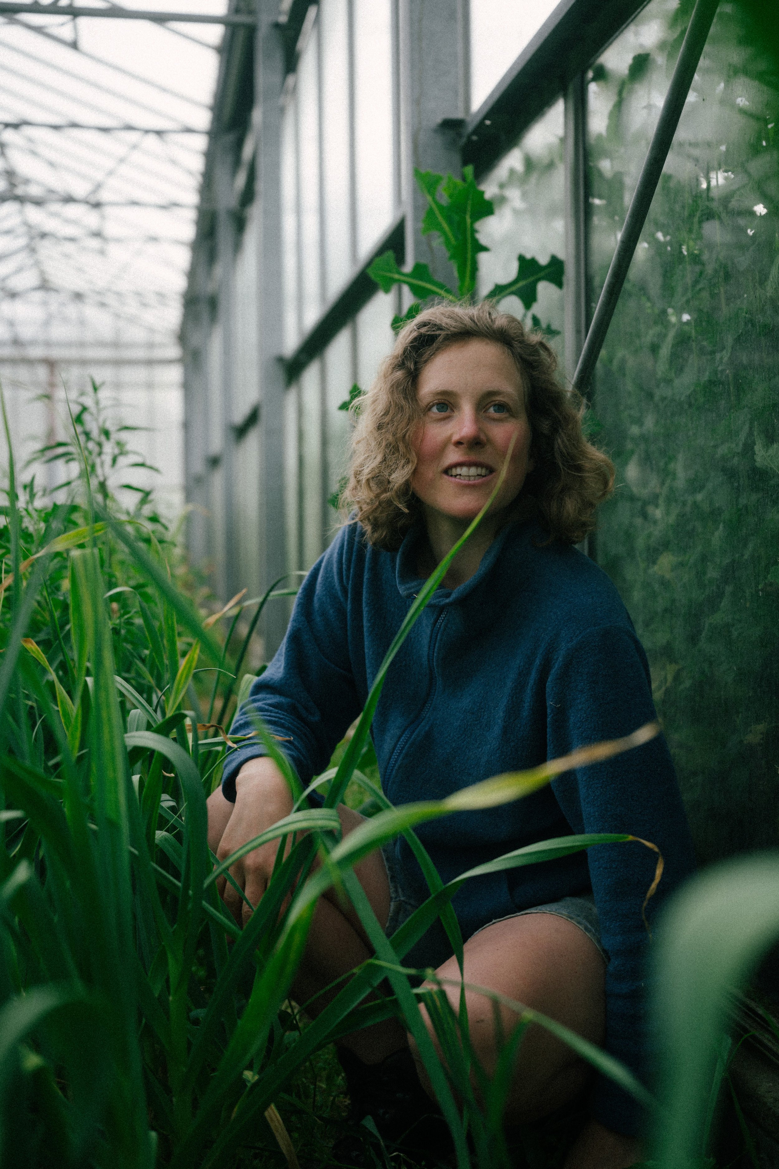 A woman with curly hair smiling while sitting among green plants inside a greenhouse.
