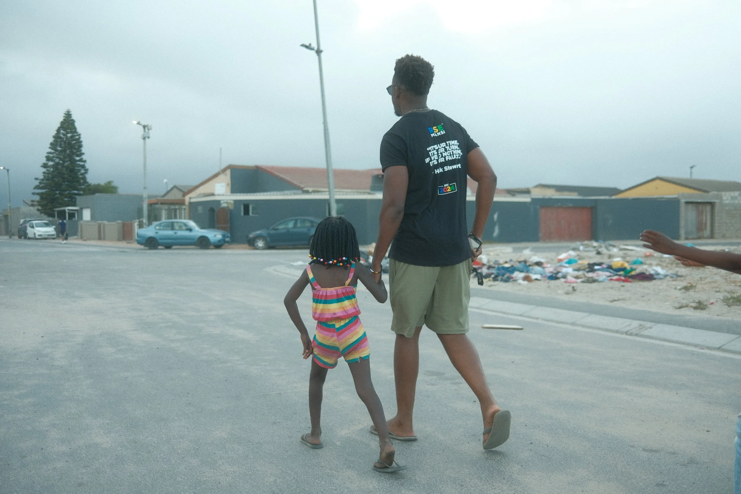 A man and a young girl walking hand in hand on a street with a pile of laundry and houses in the background.