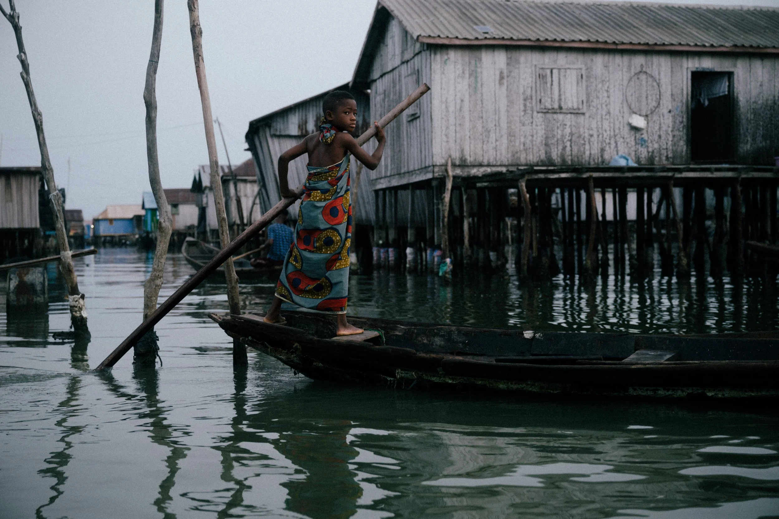 A young girl in a colorful dress stands on a small boat, holding a pole, in a water community with wooden houses on stilts.