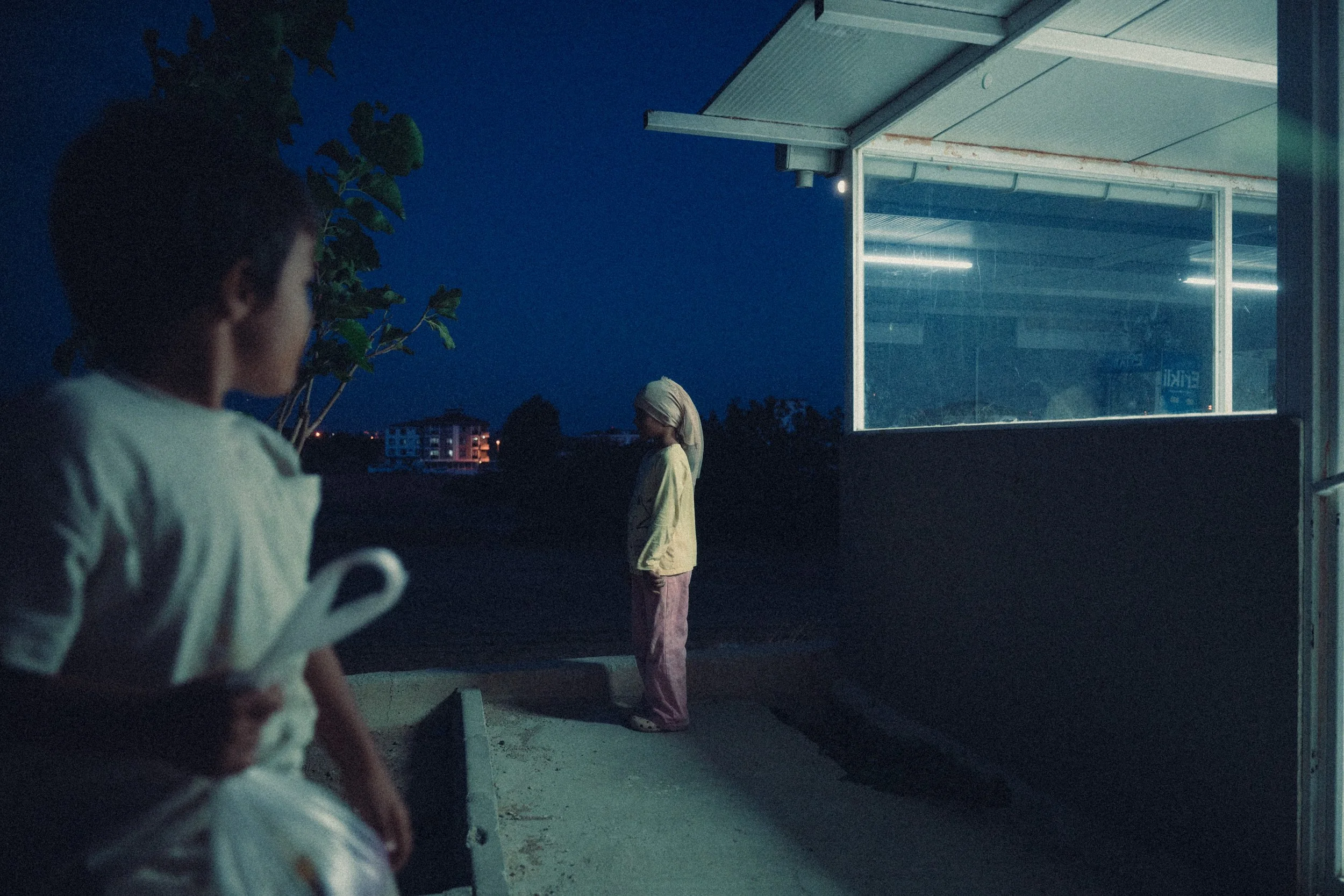 A man in the foreground holding a plastic bag and a rolled-up item, looking at a woman standing outside near a building with large windows. It is nighttime, and the scene is dimly lit.