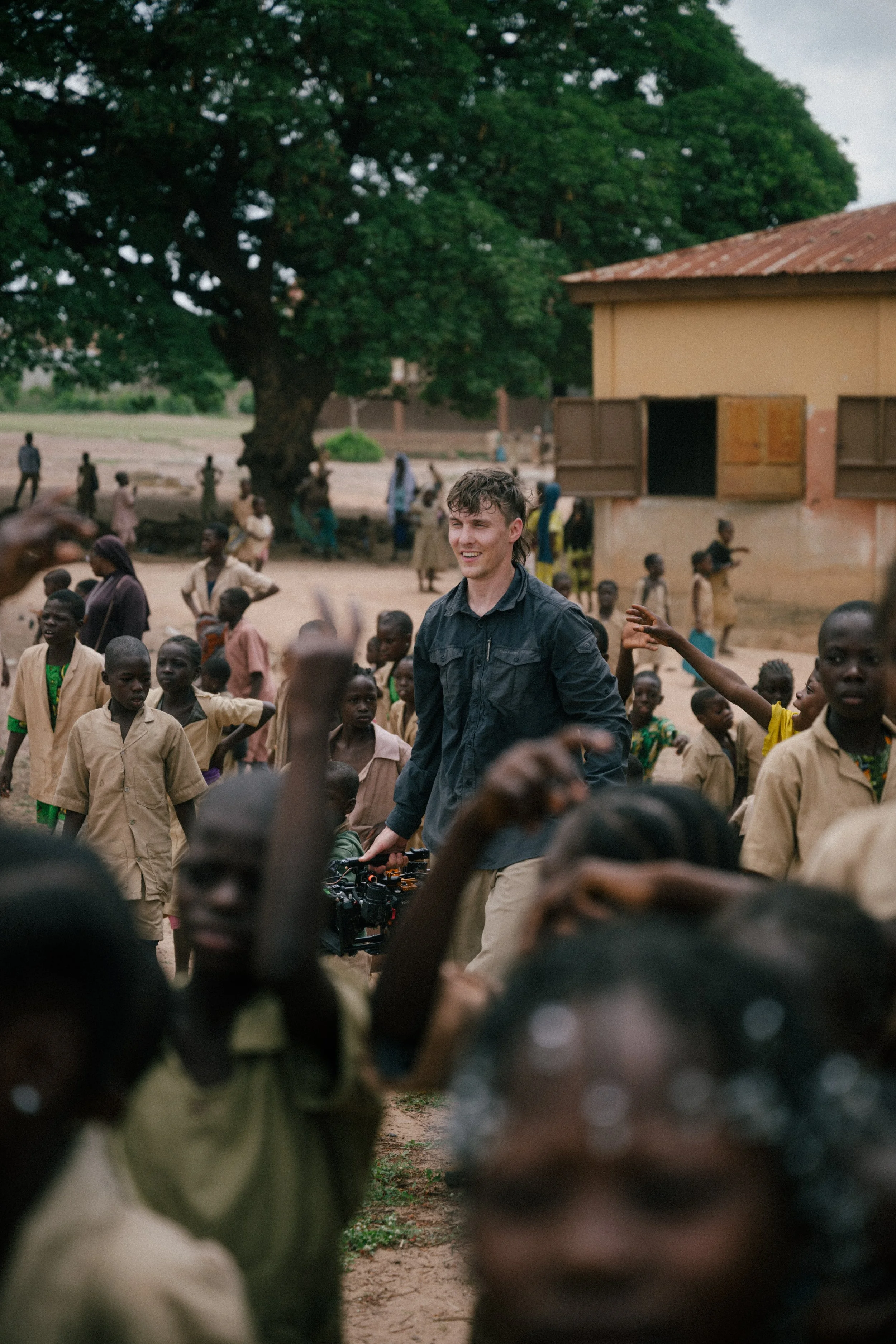 Young man with a camera smiling and walking through a crowd of children in a rural setting with trees and a building in the background.