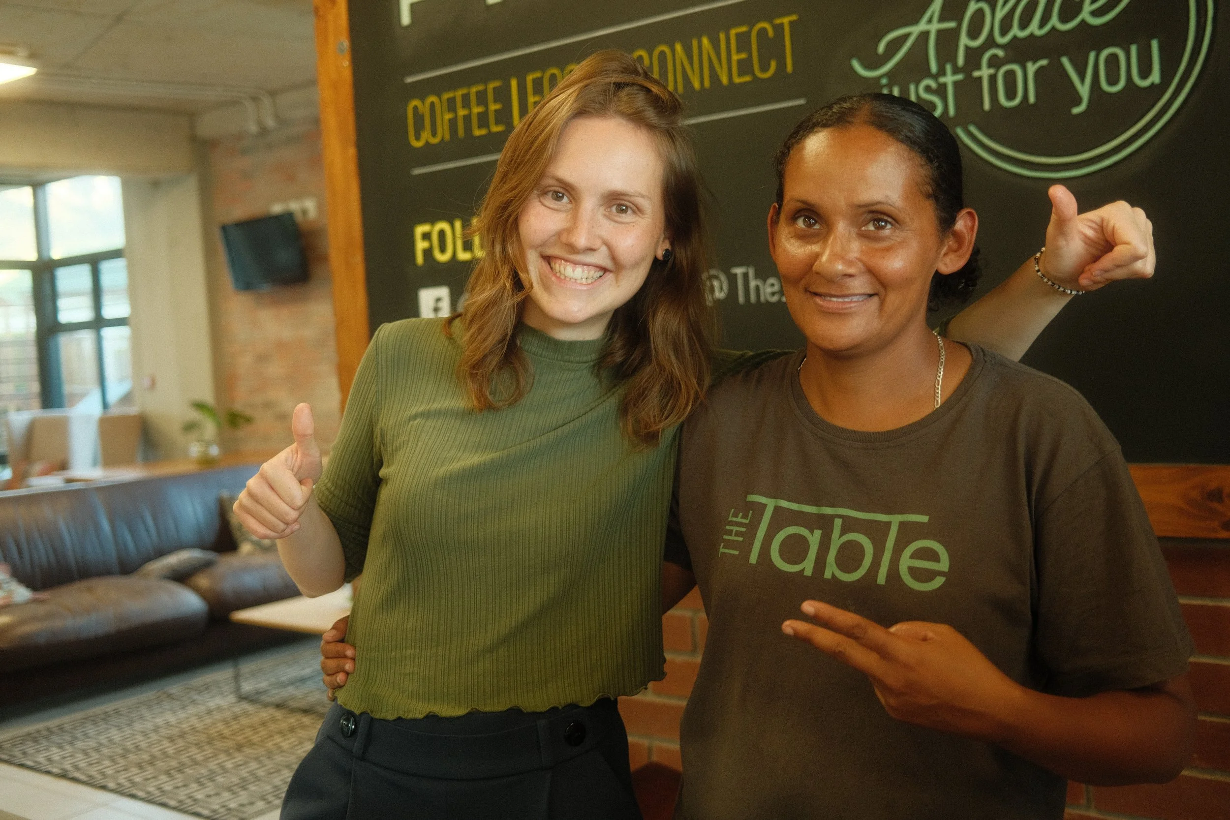 Two women smiling and posing inside a cafe with a blackboard behind them. One woman has red hair and is wearing a green top, giving a thumbs-up. The other woman has dark hair, is wearing a brown t-shirt with green text, and is making a peace sign. Th