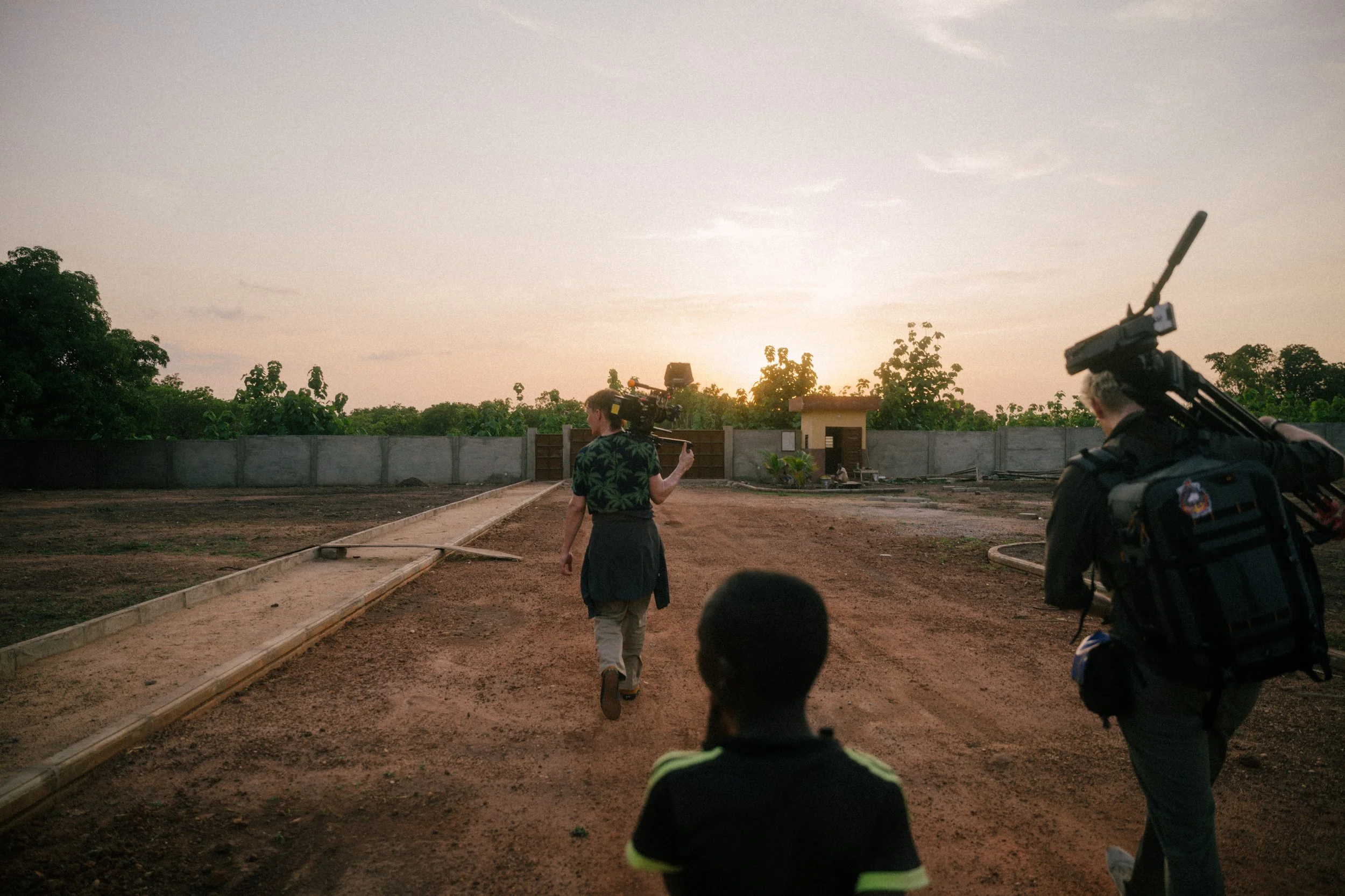 People walking on a dirt path towards a house at sunset, with one person carrying filming equipment.