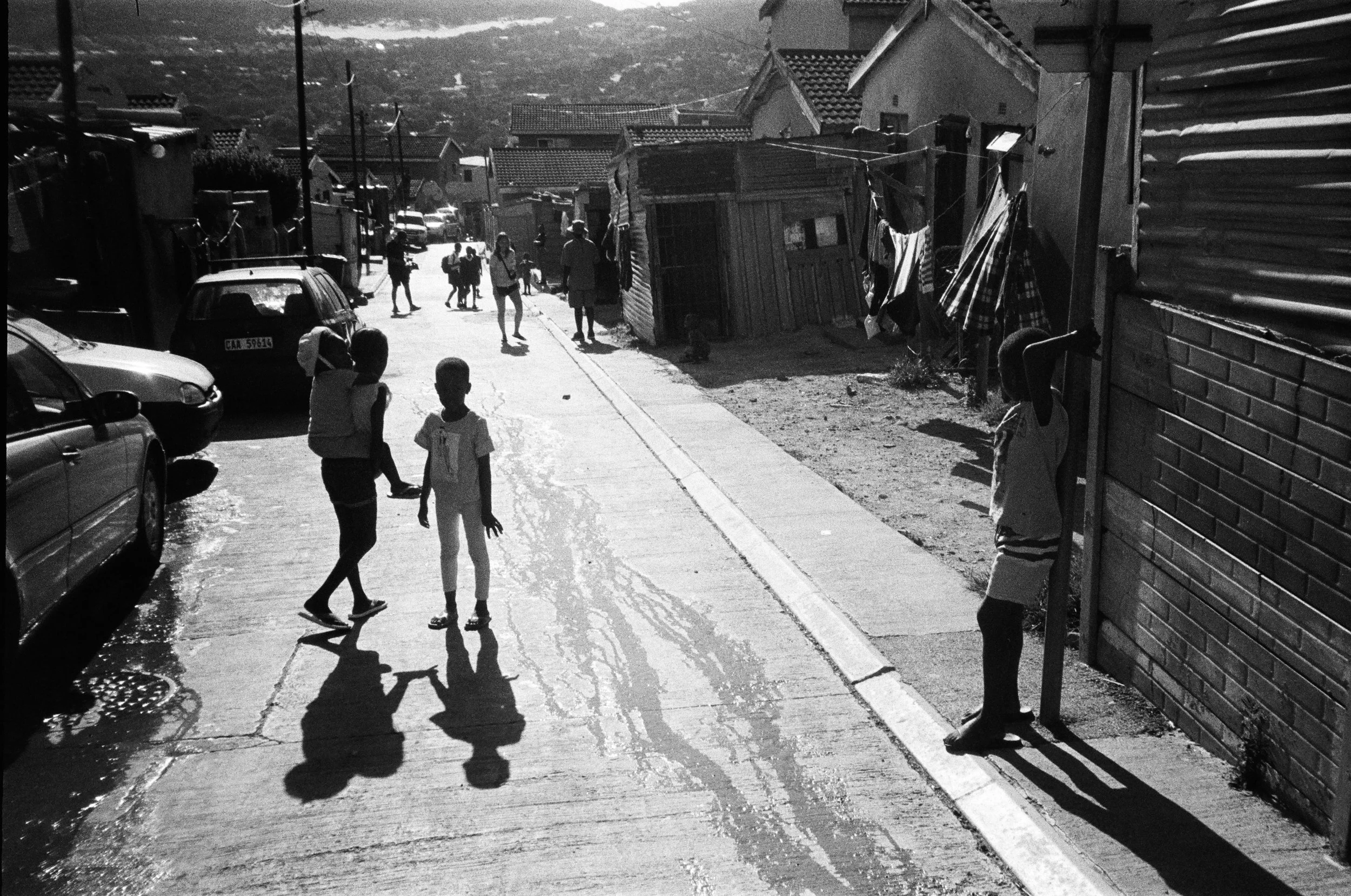 People, including children, walking on a sunlit street with parked cars and small residences, some laundry hanging outside, in a neighborhood with hills in the background.