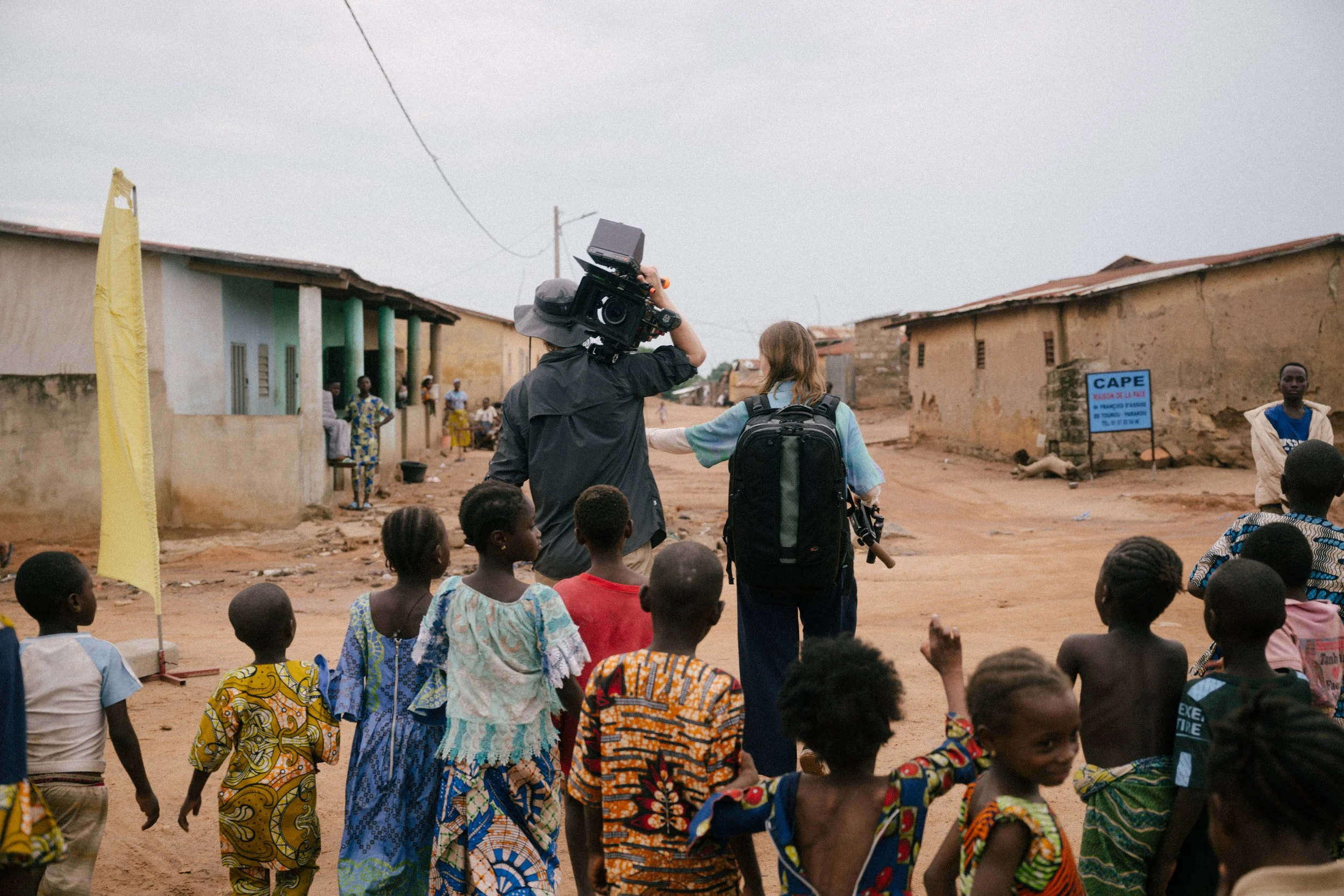 A group of children and two women, one holding a camera, walking through a village with simple houses, some adults in the background, and a sign that reads 'CAPE'.