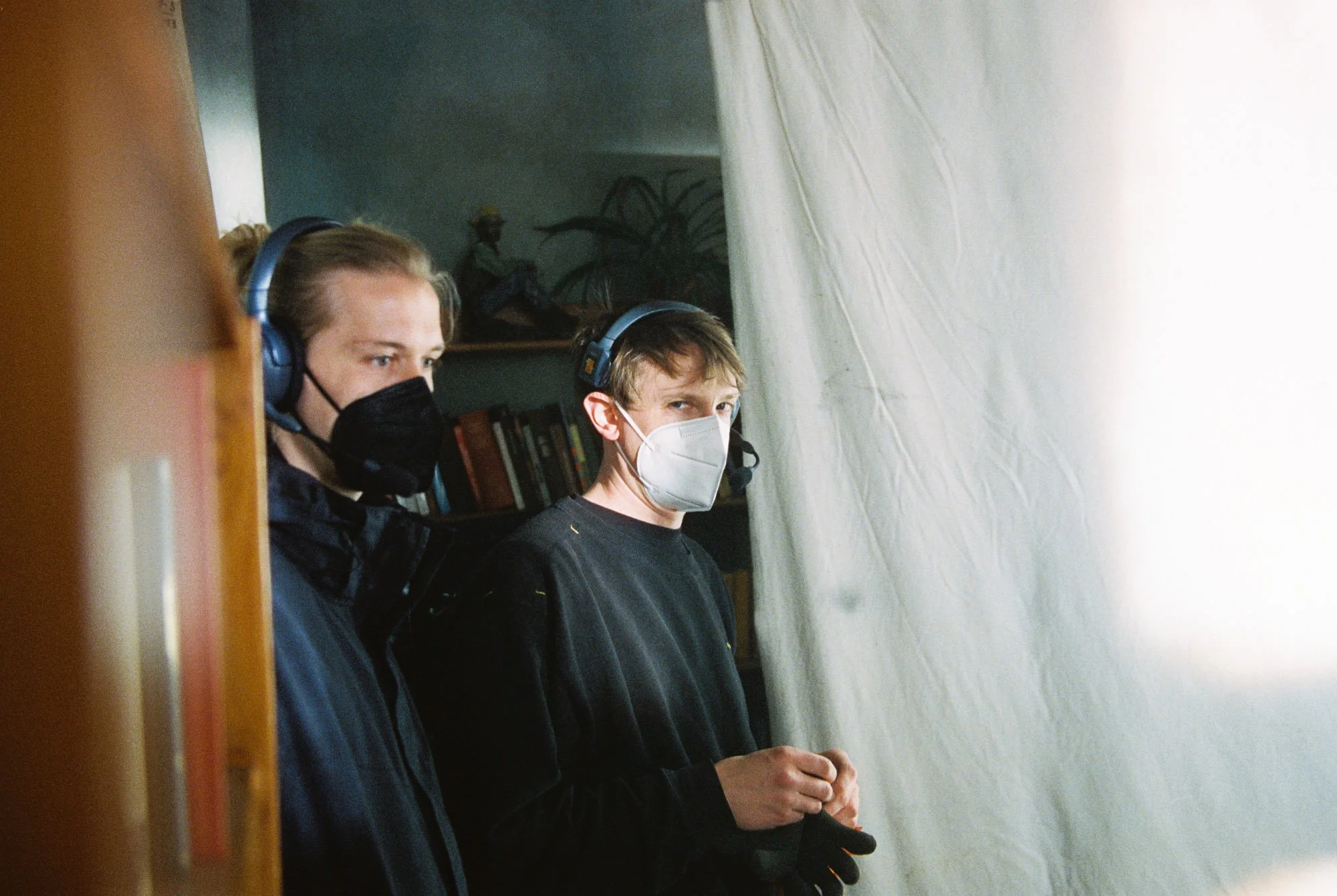 Two young men wearing face masks and headphones look intently at something off-camera. One is dressed in black and the other in dark blue, standing near a white backdrop or curtain in an indoor setting.