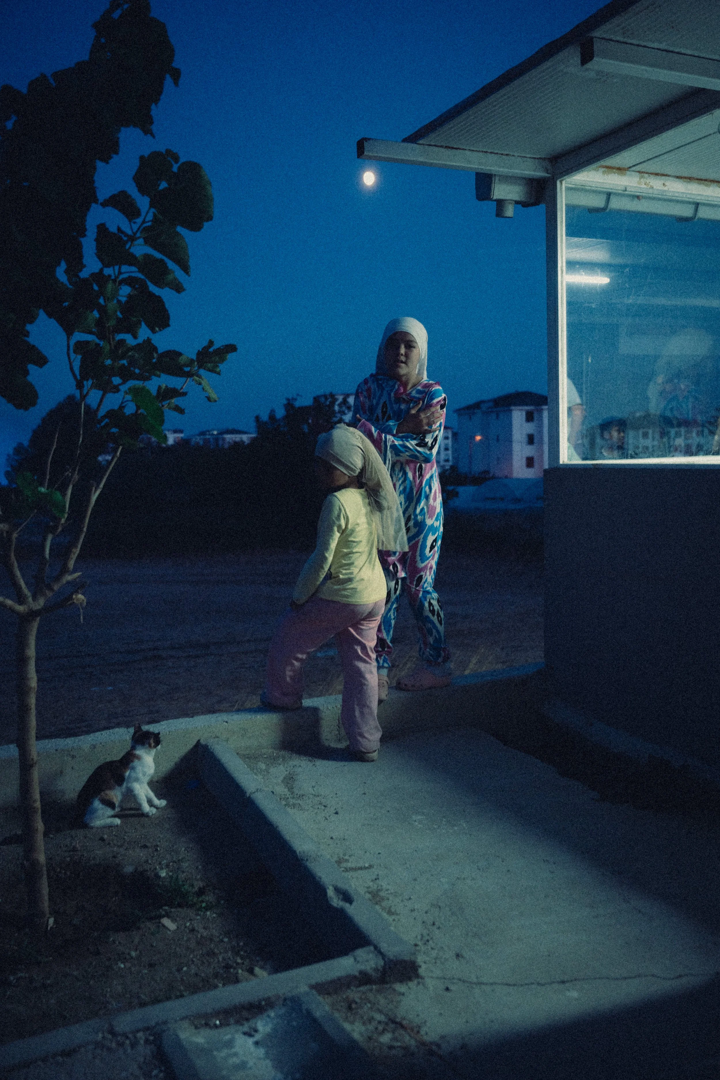 A woman and a child standing outside a small building at night, with a full moon visible in the sky. The woman is wearing a headscarf and colorful pajamas, and the child is wearing a hat and light-colored clothing. A cat is sitting on the ground near