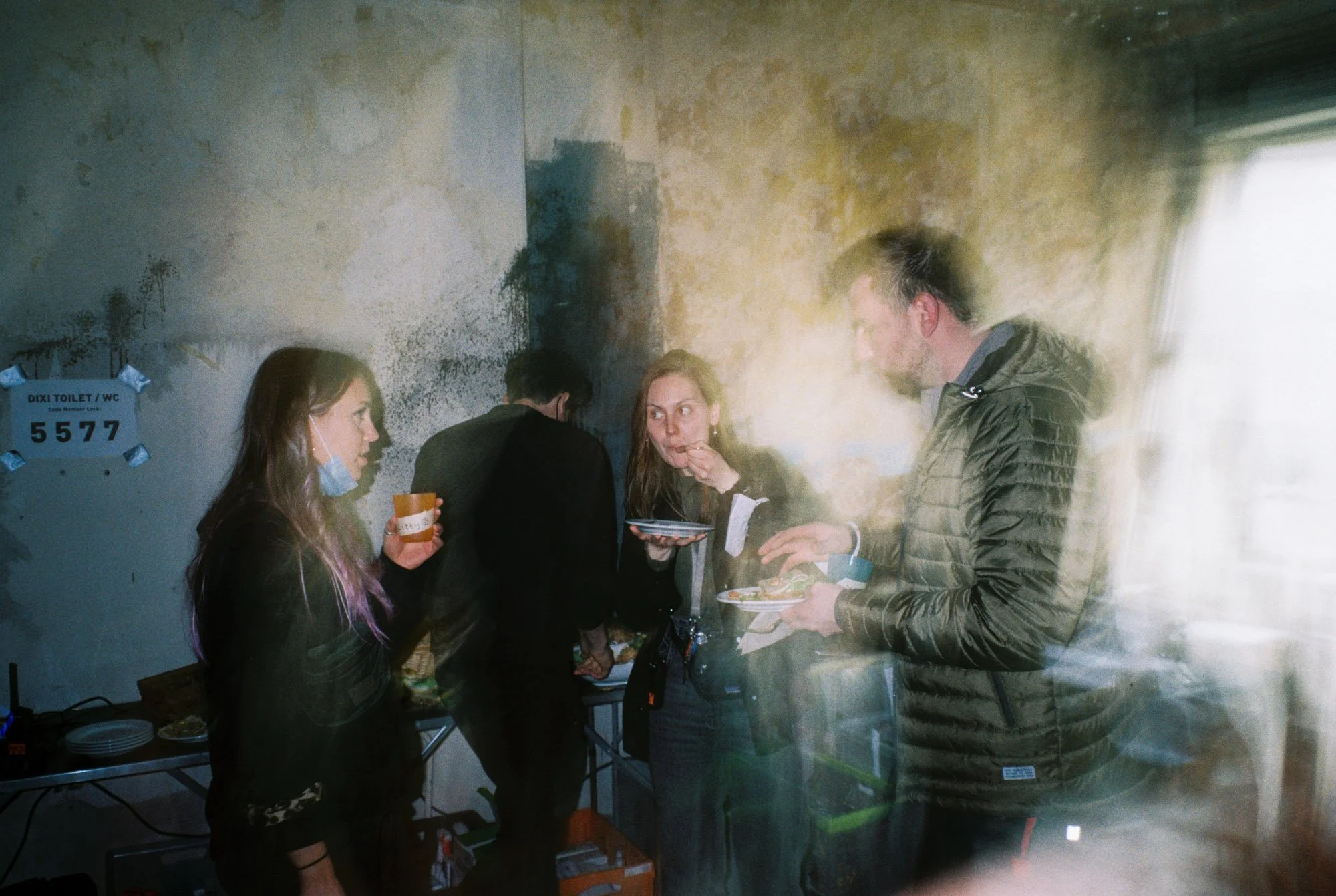 People at a gathering with food, standing near a table with dishes in a dimly lit room with a textured wall.
