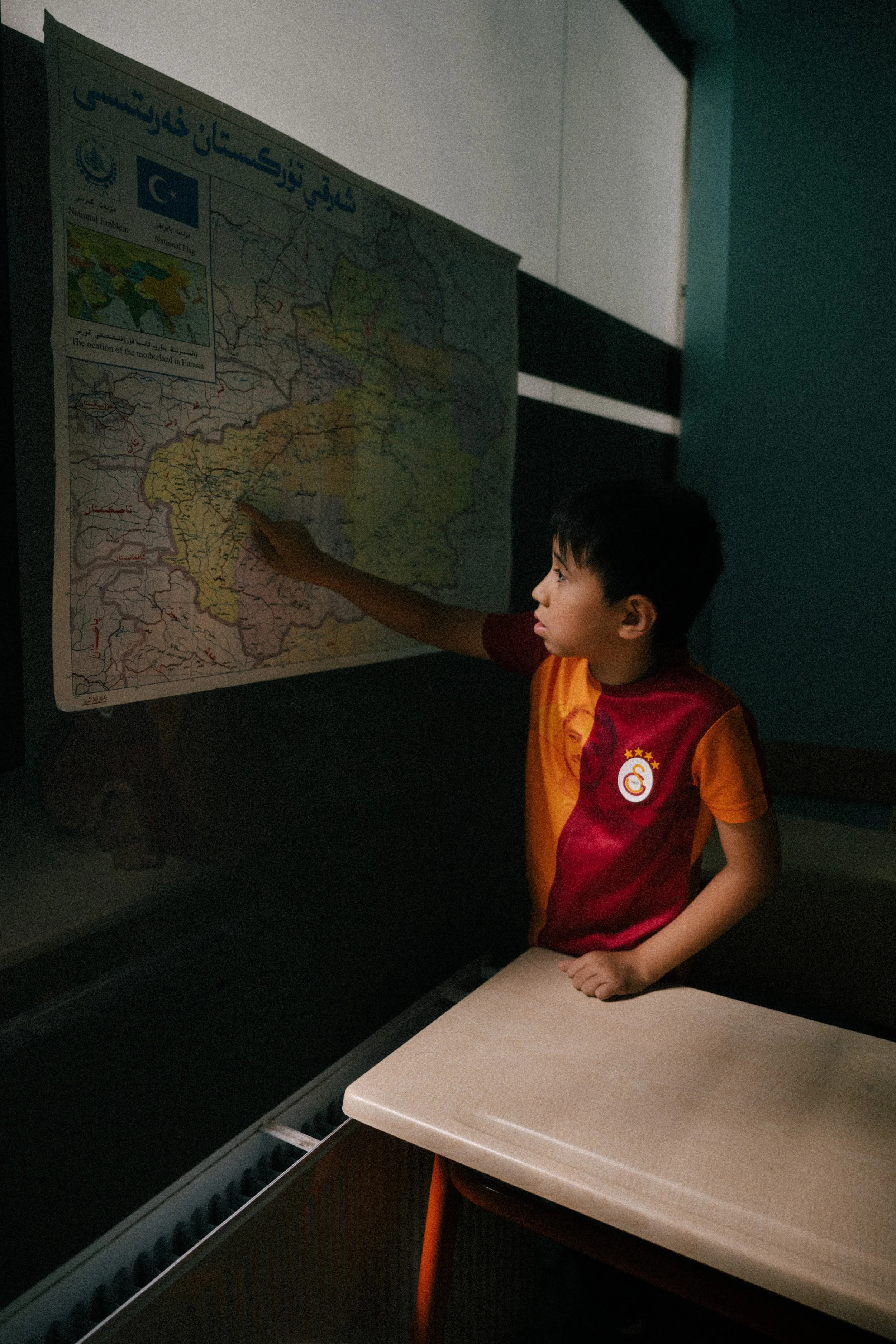 A young boy in a Galatasaray football jersey points to a map of Afghanistan on a classroom wall.