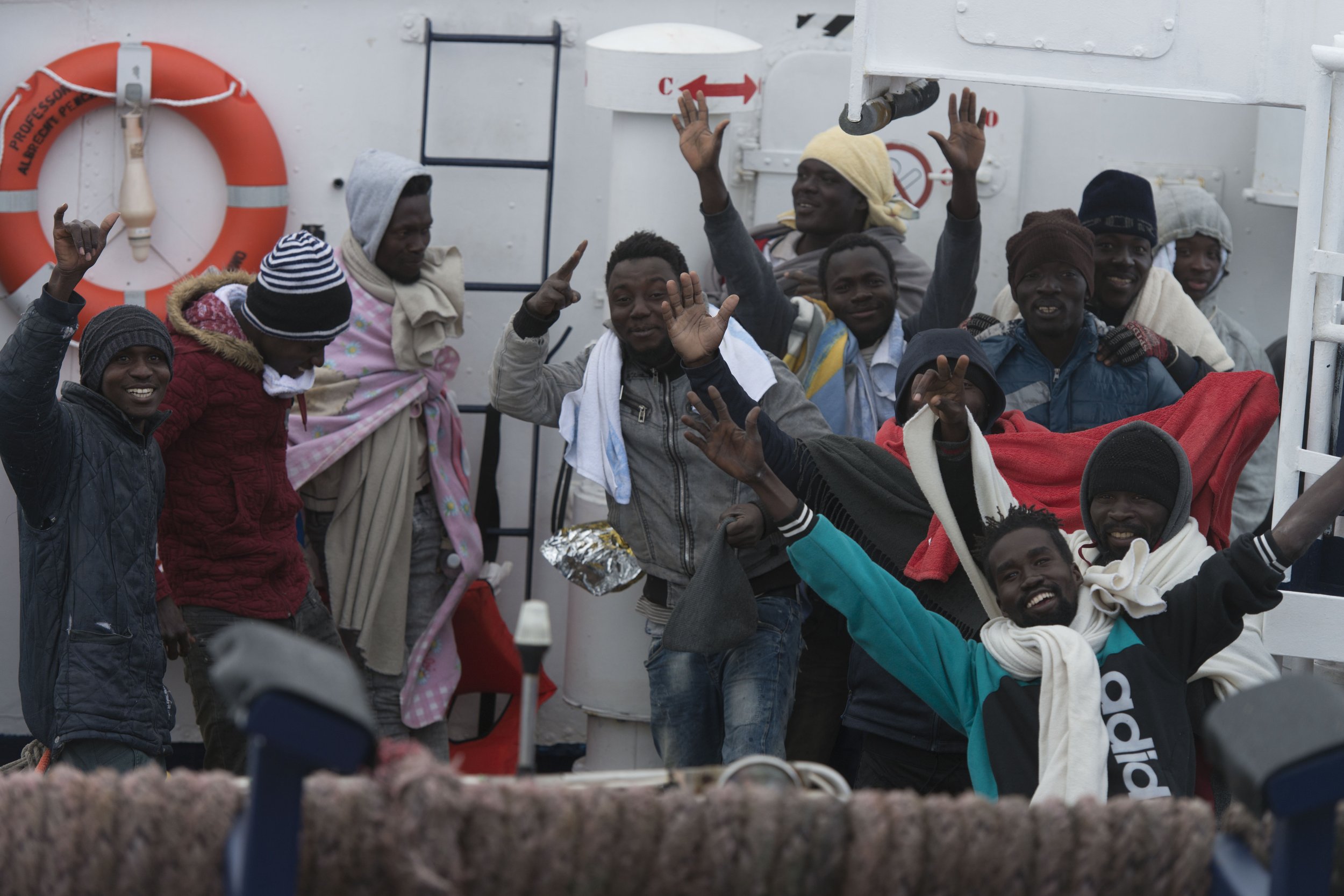 A group of men and boys aboard a boat, smiling and waving, dressed warmly with various jackets, hats, and scarves, with a life ring and safety equipment visible in the background.