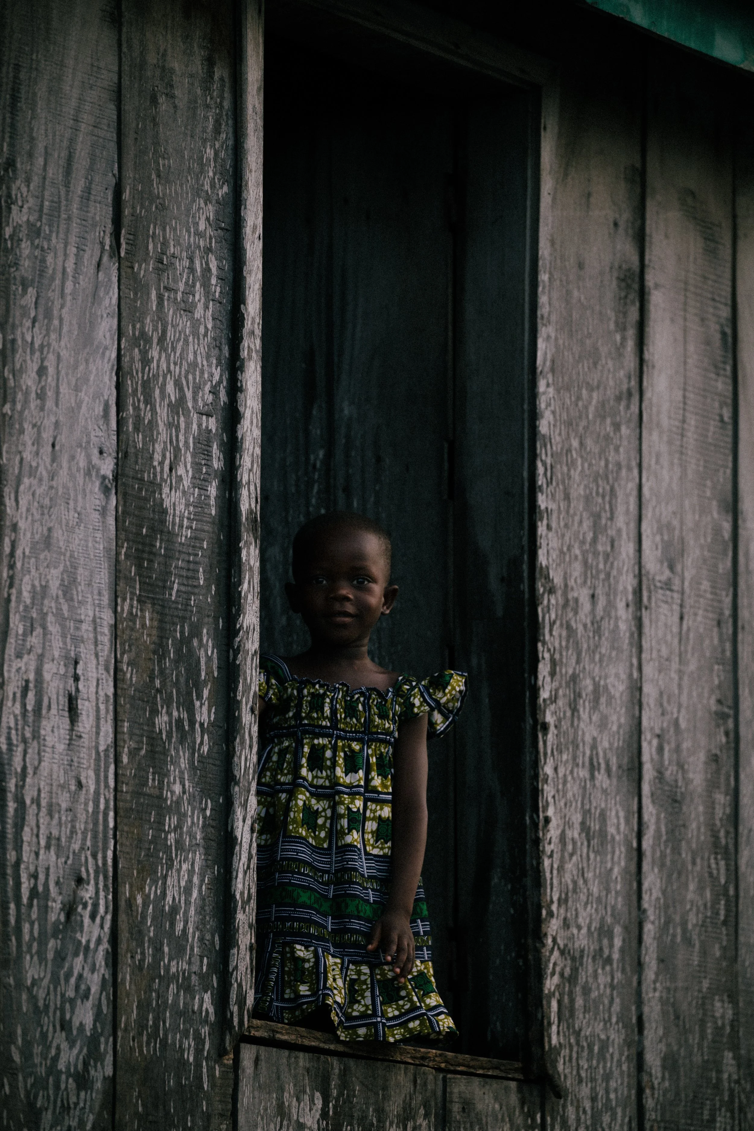 A young girl standing in a wooden doorway of a weathered house, looking out with a slight smile.
