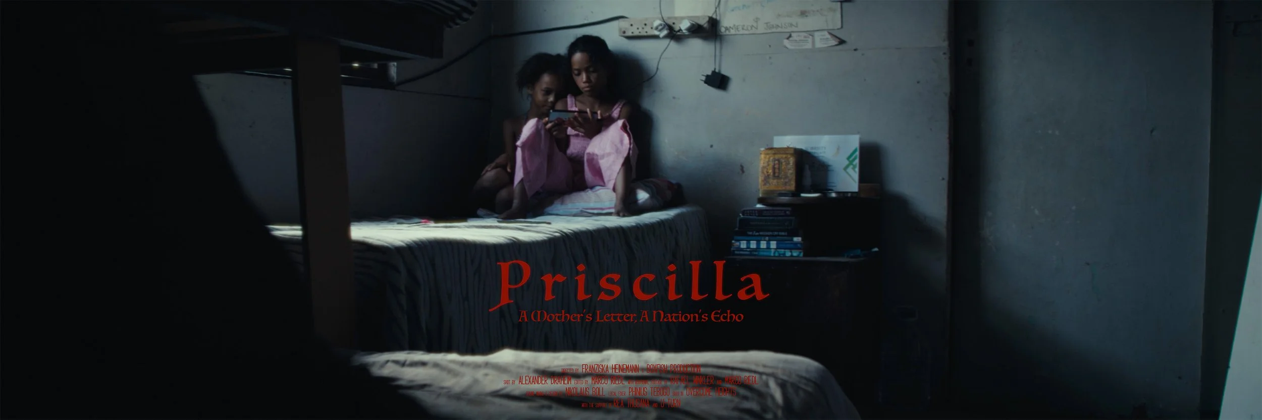 Two young girls sitting on a bed in a small, dimly lit room, looking at a mobile phone. The room has a small table with books and a box on it, and a whiteboard or wall with wires attached. The scene appears intimate and quiet, with the girls focused on the phone.