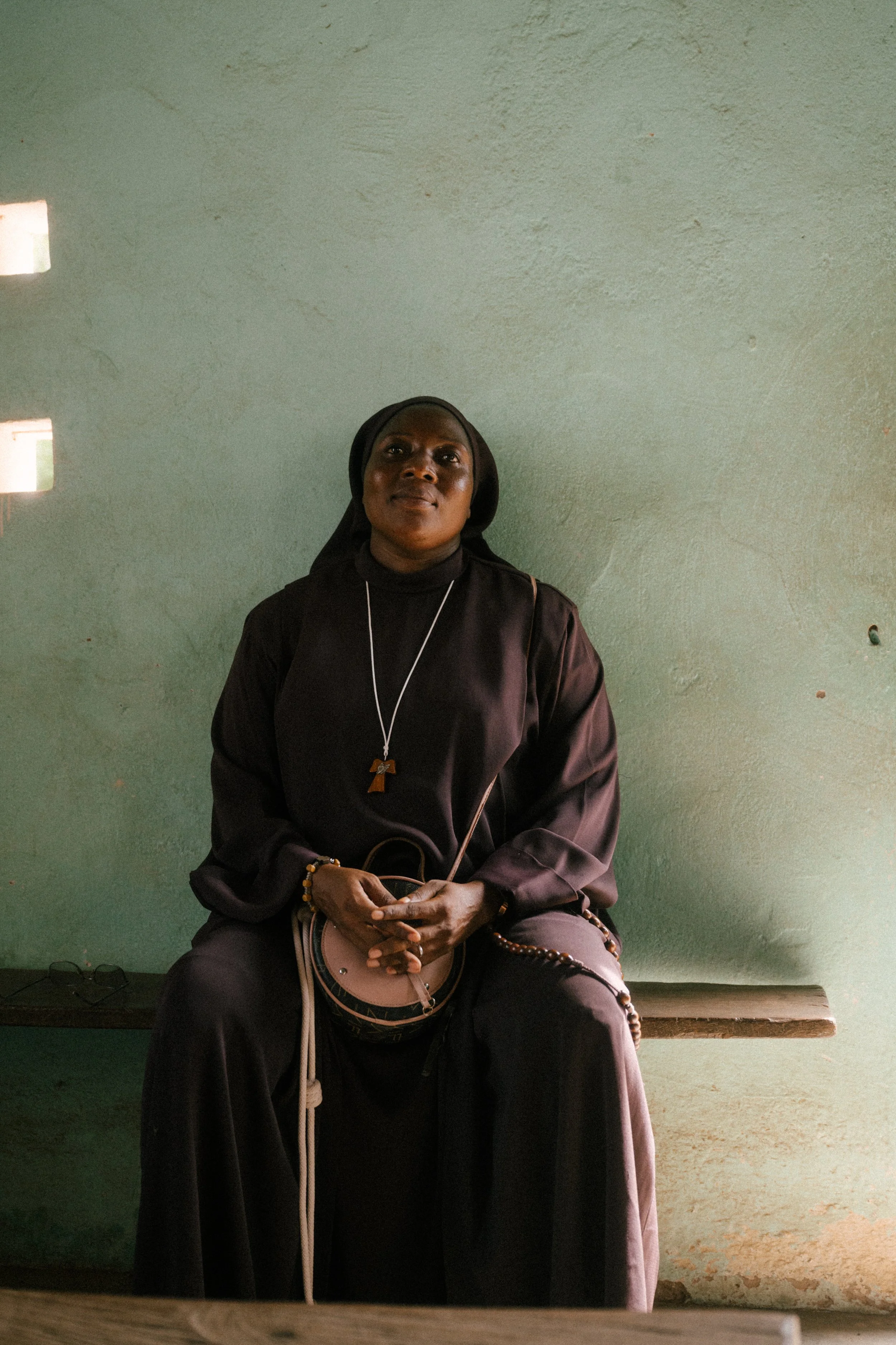 A woman dressed as a nun sitting on a wooden bench against a green wall, holding a beige purse, with her hands clasped, wearing a black habit, a cross necklace, and a bracelet.