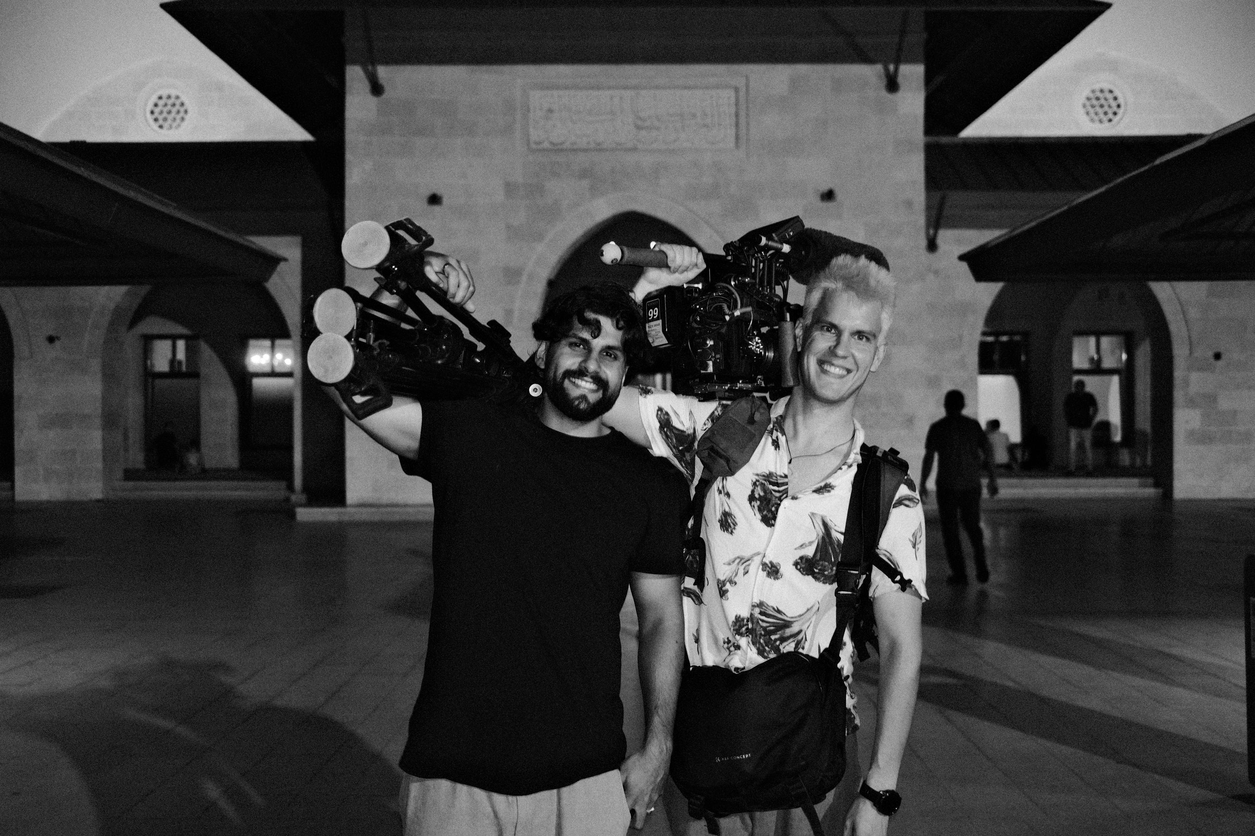 Two men smiling at night, one carrying a large camera crane on their shoulder, in front of a building with arched entryways and a brick facade.