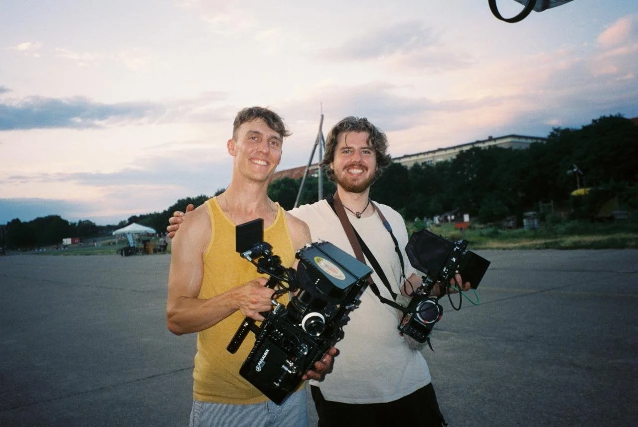 Two men standing outdoors on a paved area holding camera equipment, smiling at the camera. One wears a yellow tank top, the other a white t-shirt. The background features trees, a cloudy sky, and distant buildings at sunset.