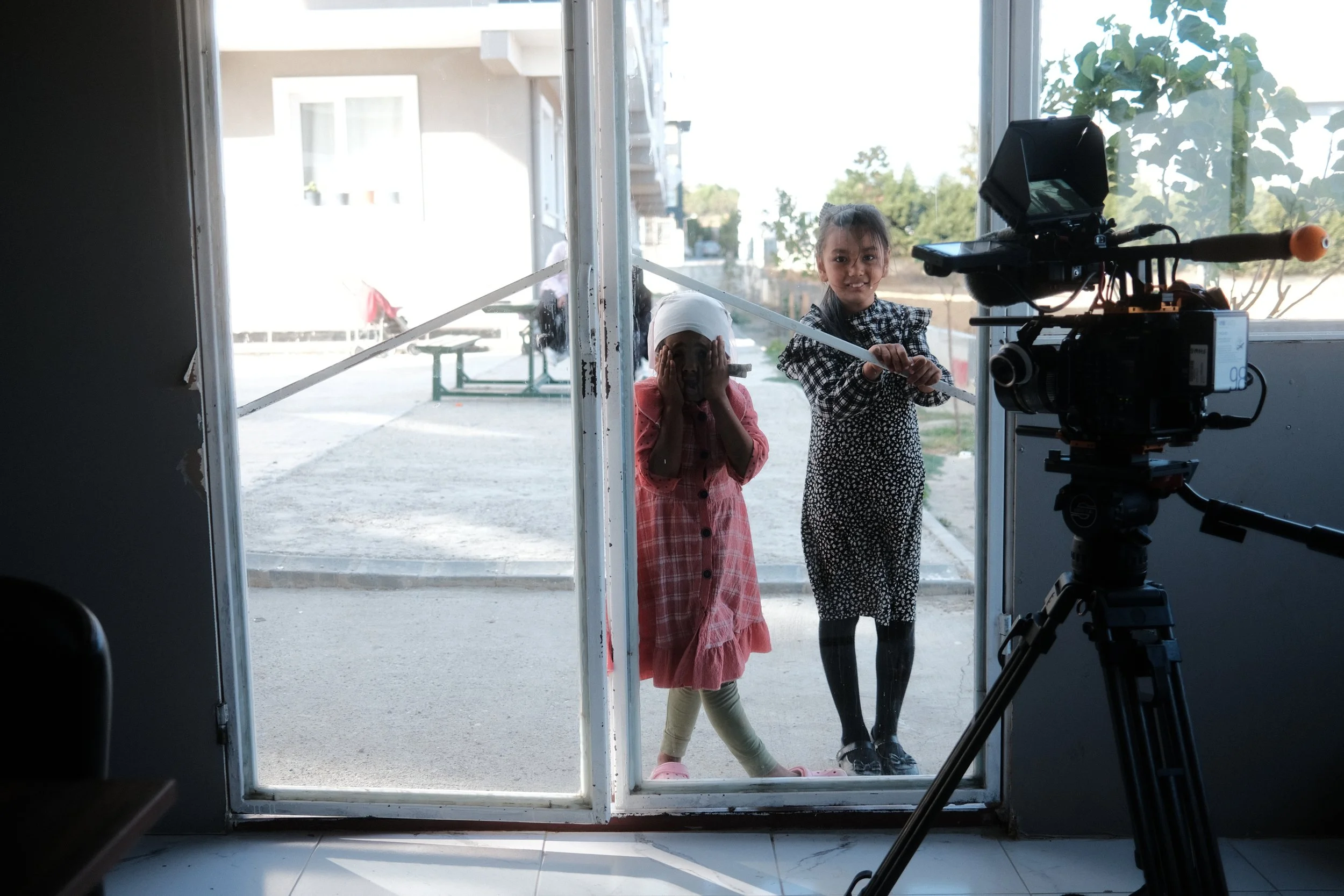 Two children inside a building looking through glass door at a camera outside. One girl is wearing a pink dress and has a napkin wrapped around her head, holding her face with her hands. The other girl is wearing a black and white dress, holding a lo