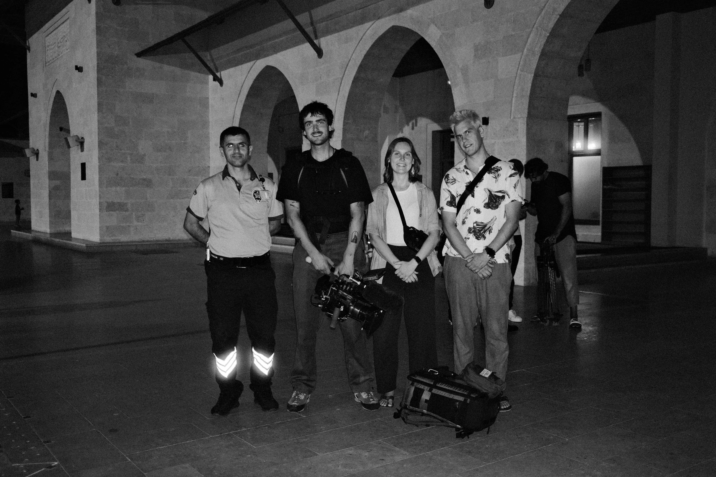 Four people stand together in a building with stone arches, two others are in the background, one adjusting equipment.