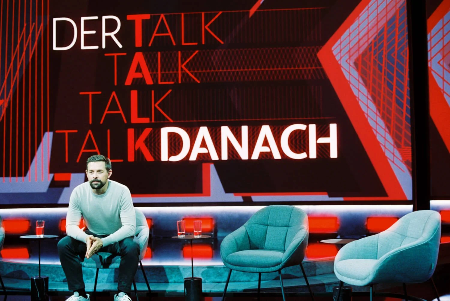 A man with a beard and a white sweater sitting on a stage in front of a large red and black backdrop with the words 'DER TALK' and 'TALK' repeatedly written, and the word 'DANACH' illuminated at the bottom.