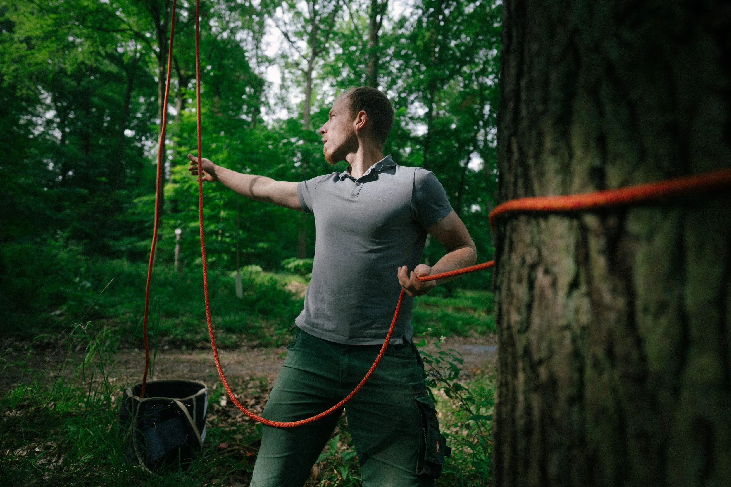 A man stands in a forest, holding orange climbing ropes and wearing hiking gear, preparing for outdoor activity.