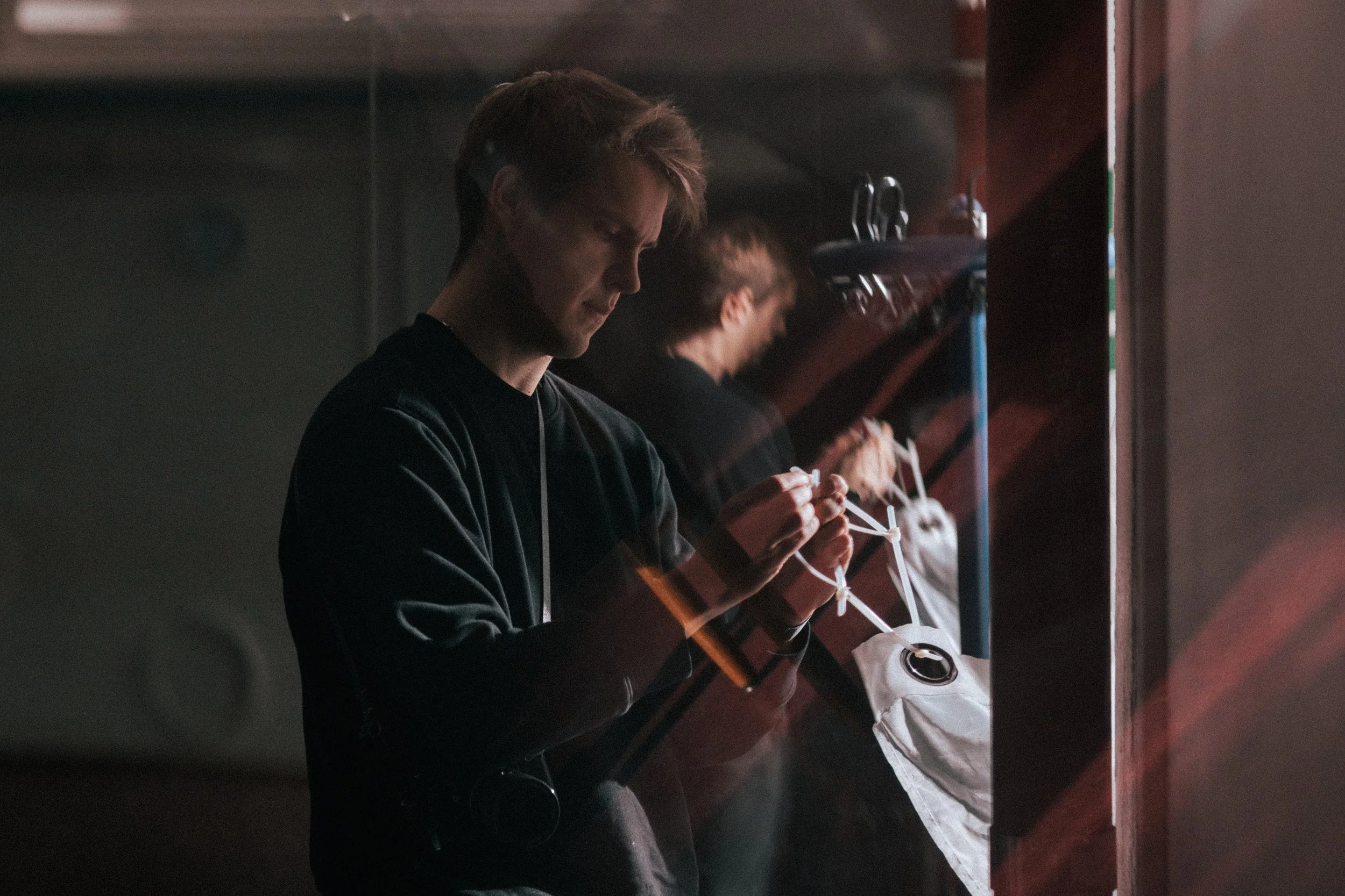 A young man with brown hair is standing at a counter, looking down at and holding a face mask with both hands. There are two other people in the background also holding face masks. The setting appears to be indoors with dim lighting.