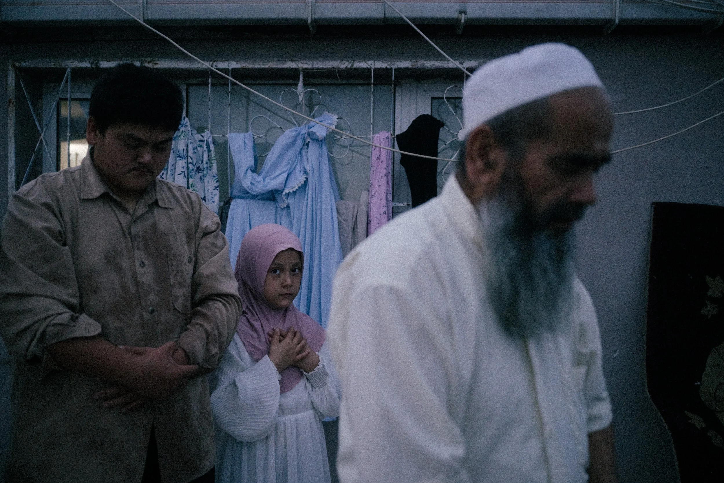 A man with a white beard and wearing a white cap and shirt is in the foreground with his head bowed. Behind him, a young girl in a pink headscarf and white dress looks directly at the camera with hands clasped. Next to her, a boy with dark hair in a 