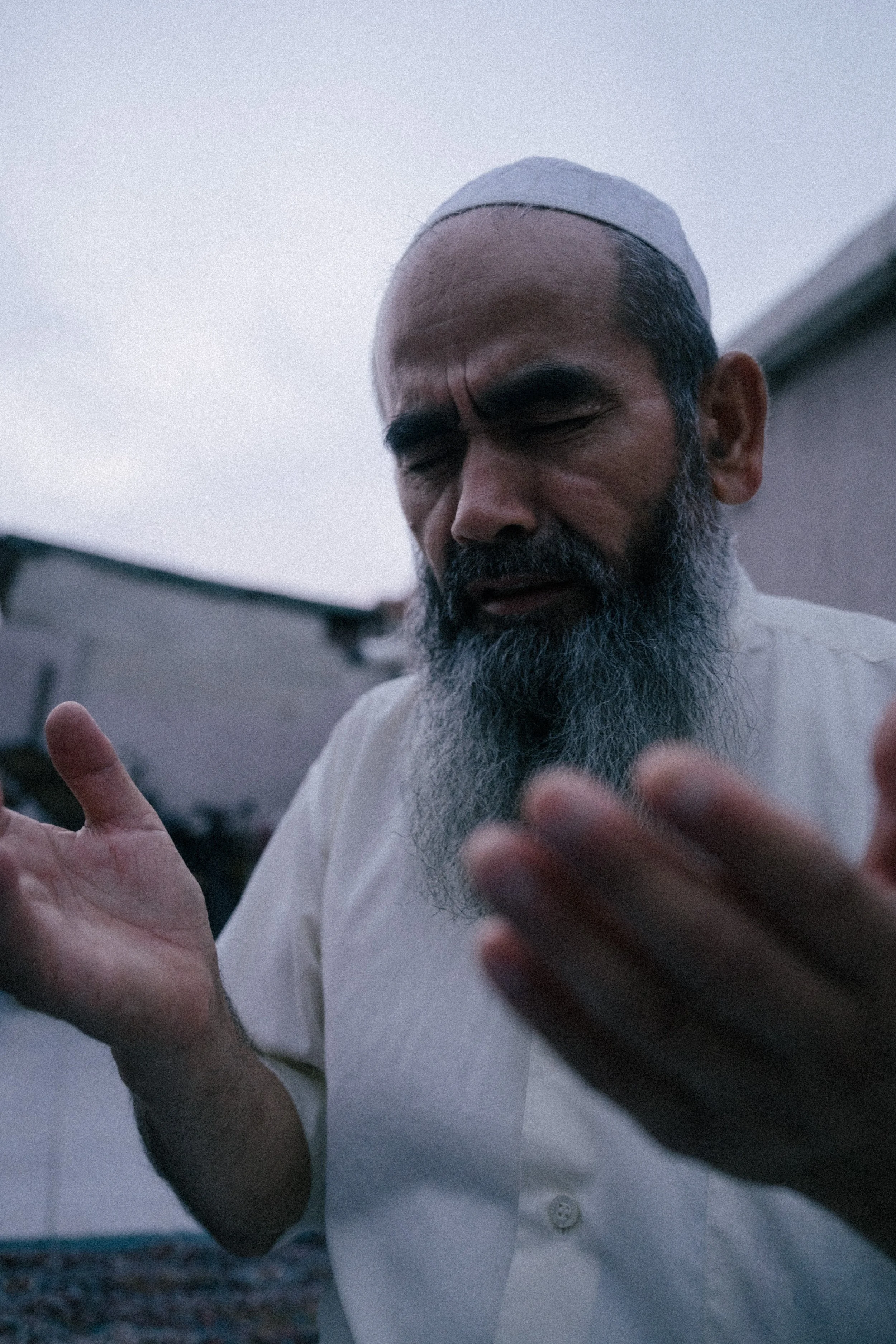A middle-aged man with a gray beard, wearing a white cap and white shirt, with his eyes closed, appears to be praying or in deep thought outdoors.