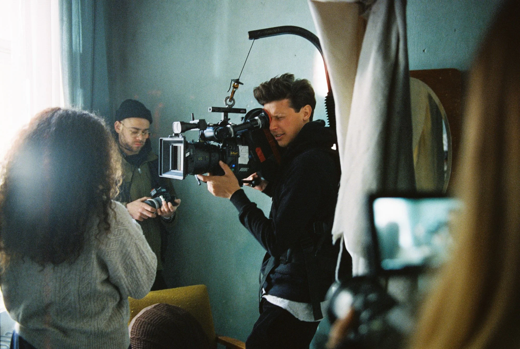 Filming scene with a man operating a professional video camera indoors, surrounded by crew members, some holding equipment, near a wall with a mirror.