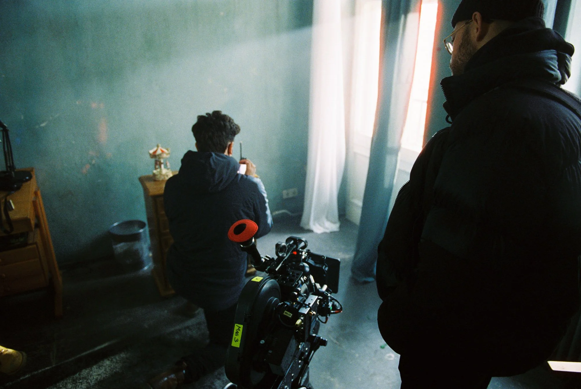 Two men behind the camera, one sitting on a chair and the other standing, filming or taking a photo of a person sitting at a small table near a window with white curtains. The person at the table has short dark hair and is holding a small object, pos