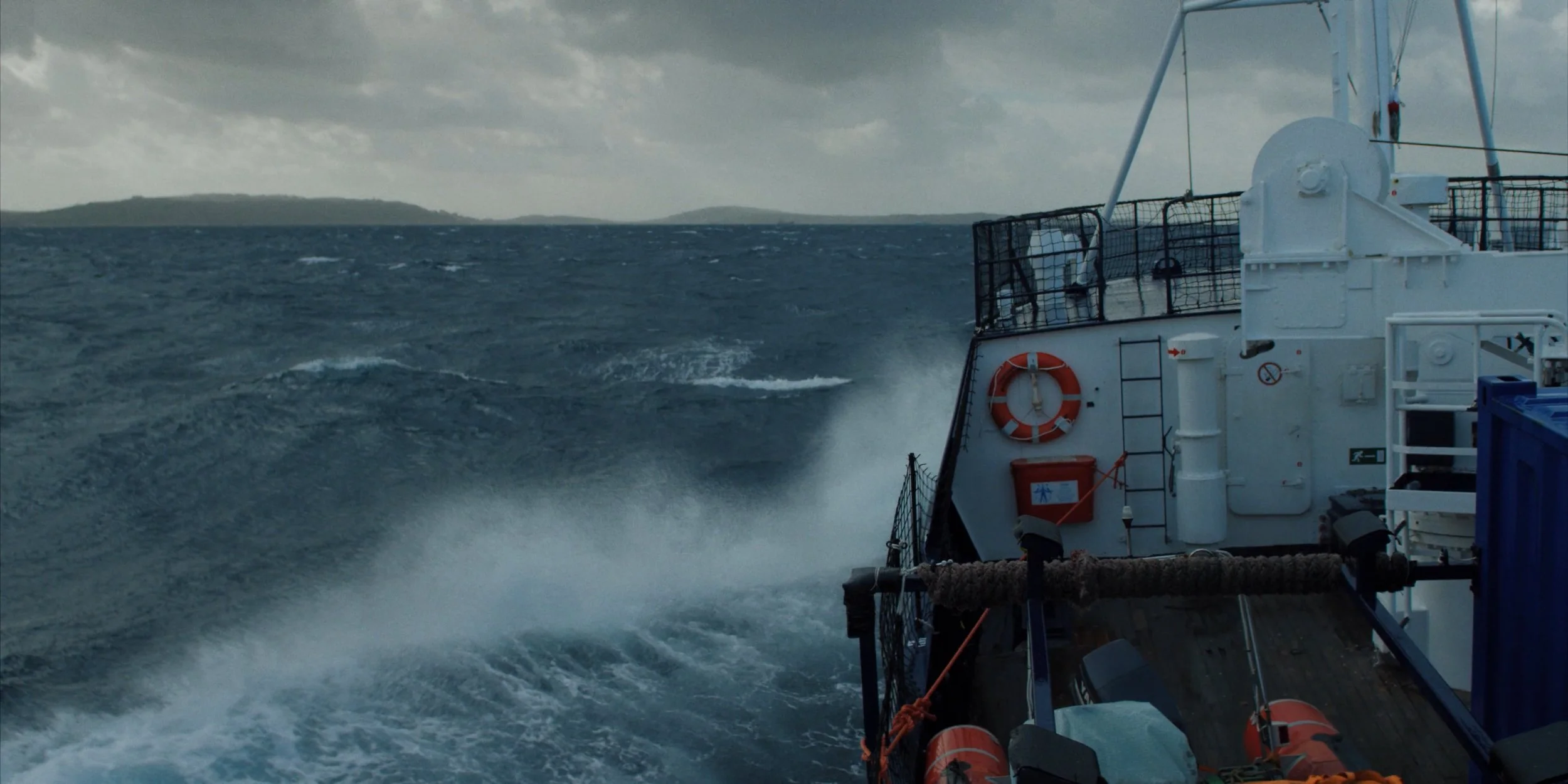 View from a boat on choppy ocean waters showing part of the boat with safety equipment, ropes, and fencing, with distant landmass under cloudy sky.