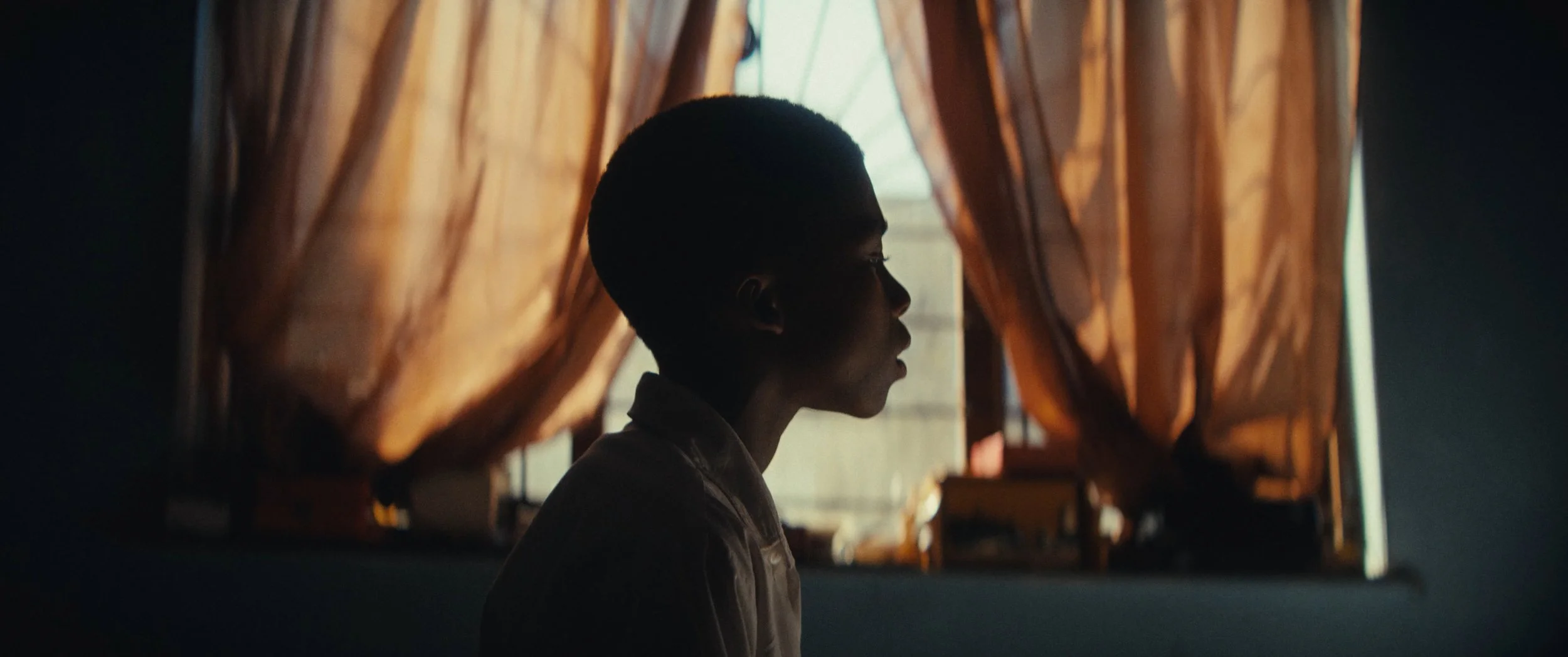 A silhouette of a young person with short hair sitting by a window with orange curtains, with soft natural light illuminating their profile.