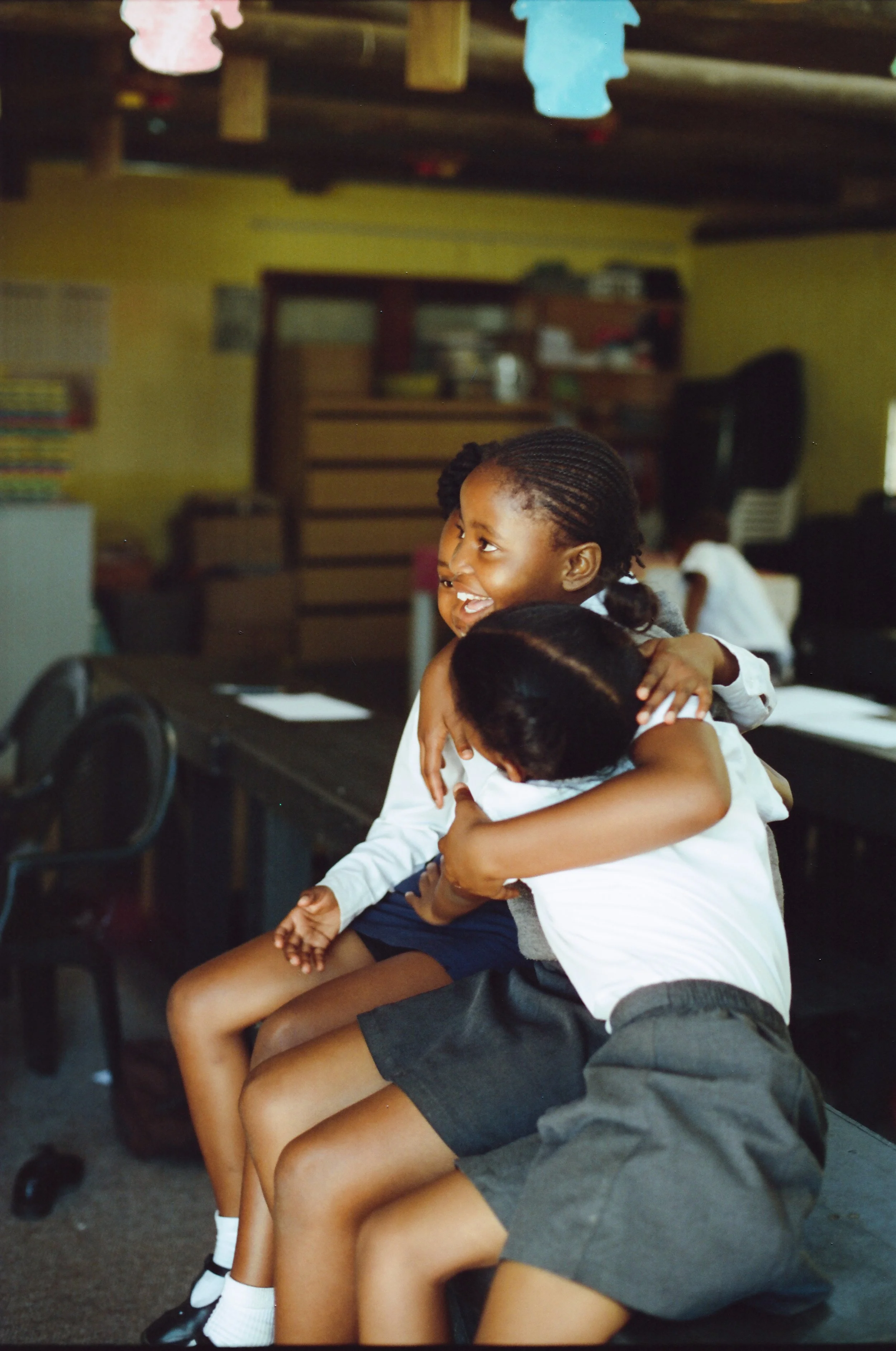Two young girls hugging and smiling in a classroom setting.
