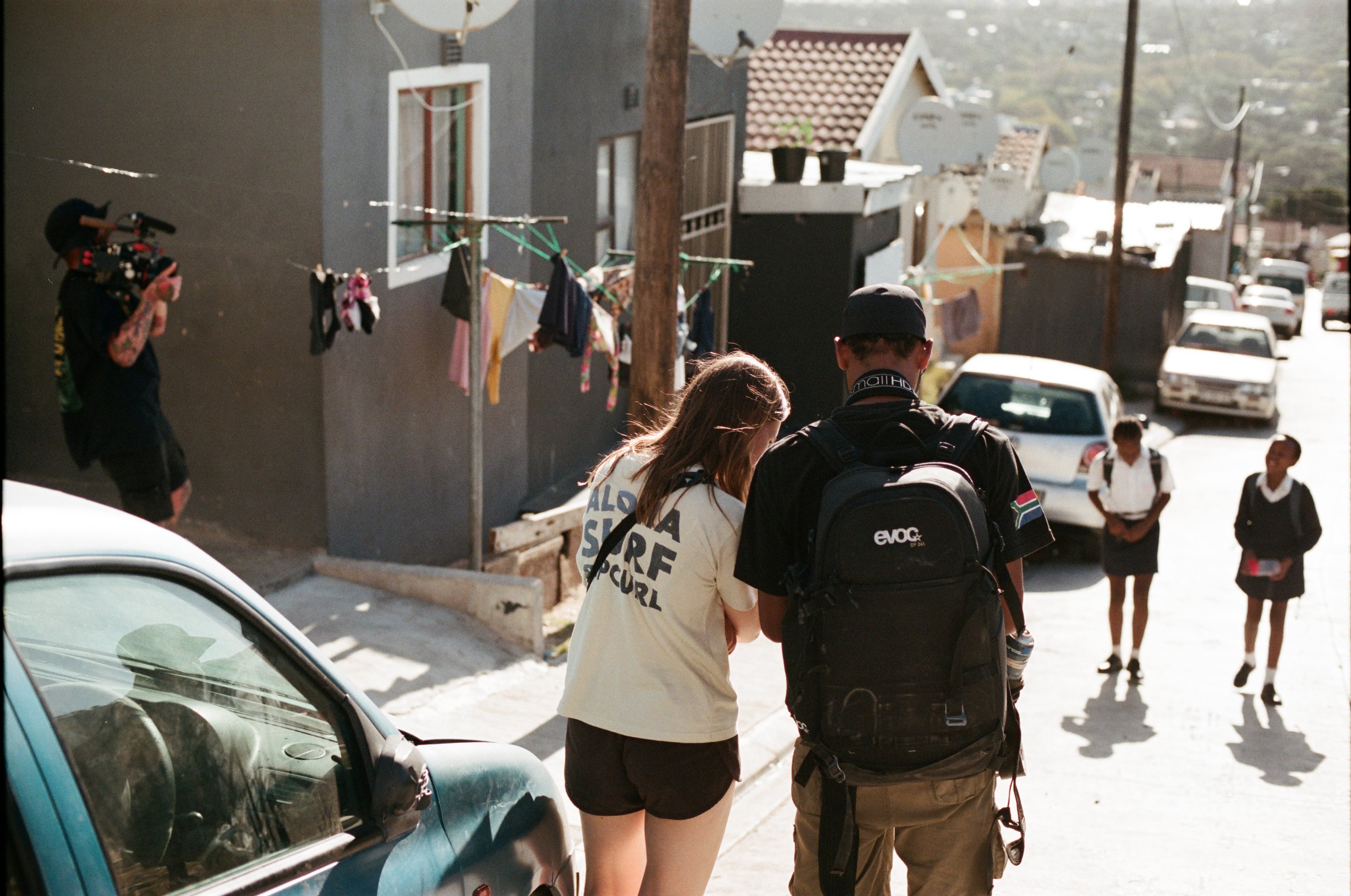 Two young people, a girl and a boy with a backpack, are walking together on a sunny street, viewed from behind. The girl has long hair and wears a white T-shirt and shorts. The boy wears a black cap and shorts. In the background, three schoolgirls in