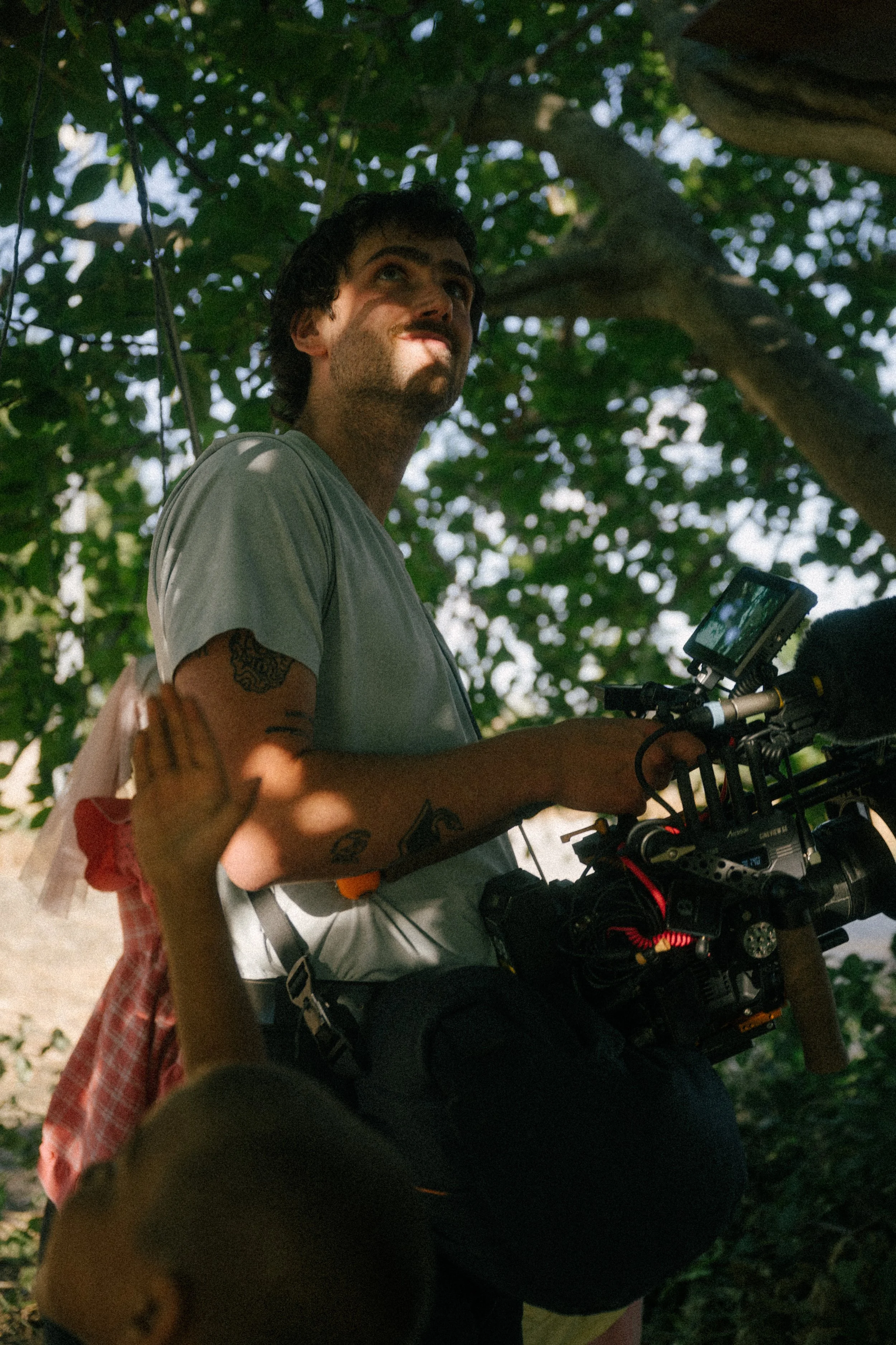 A man with tattoos standing outdoors among green leafy trees, holding a professional camera with a mounted monitor.