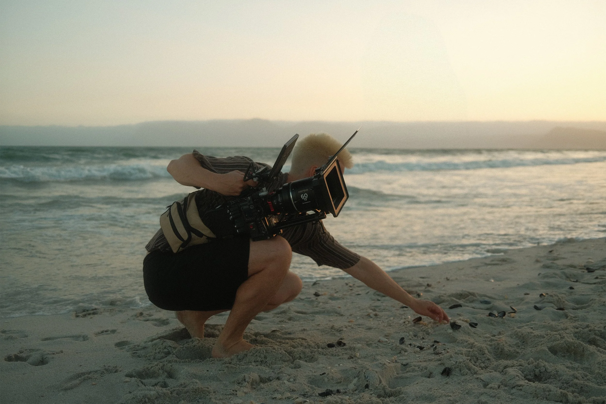A man with blonde hair is crouching on a beach, adjusting shells or small objects in the sand, with a video camera on his shoulder, during sunset.