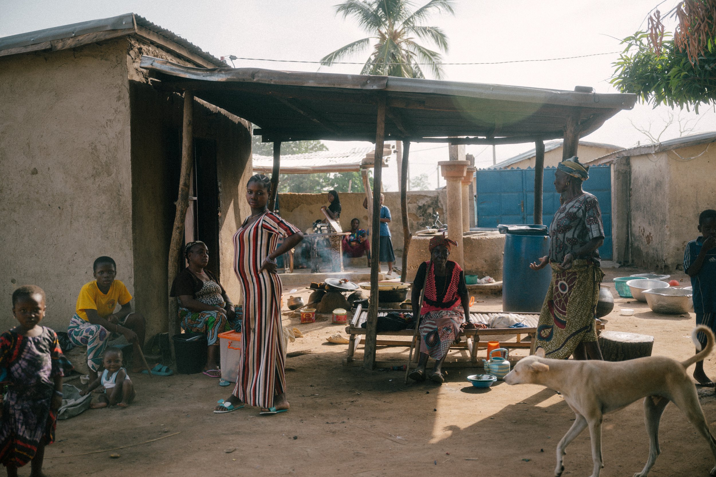 A group of people, mostly women and children, gather in an outdoor village setting, with some sitting and others standing near a simple shelter, a dog walking in the foreground, and palm trees in the background.