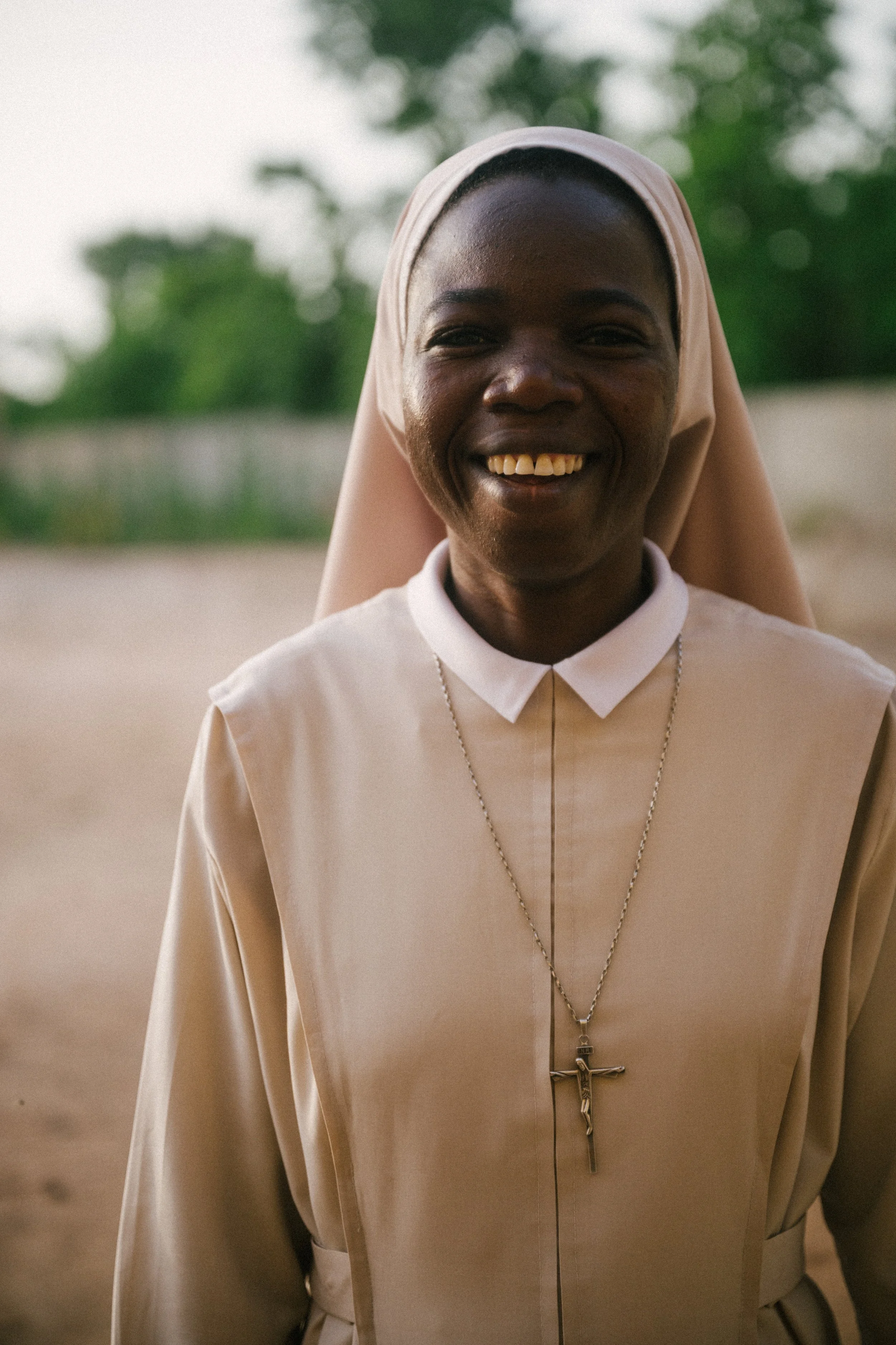 A smiling woman dressed as a nun outdoors, wearing a beige habit and a crucifix necklace.