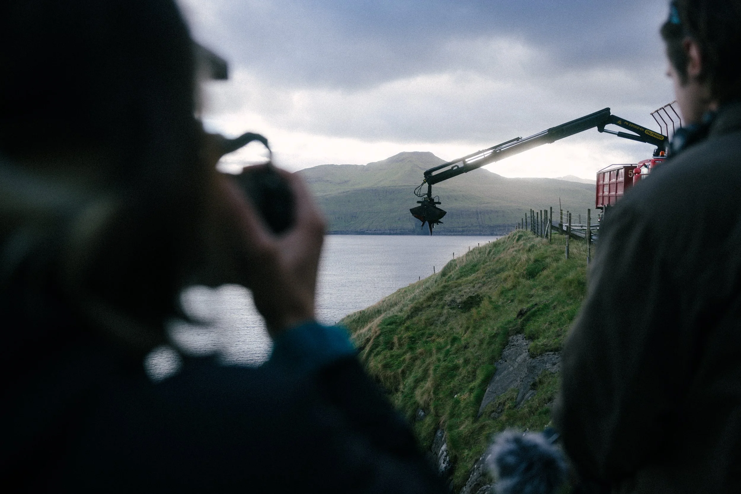 Two people are filming a landscape with a boat crane over a body of water, with hills and mountains in the background, on a cloudy day.