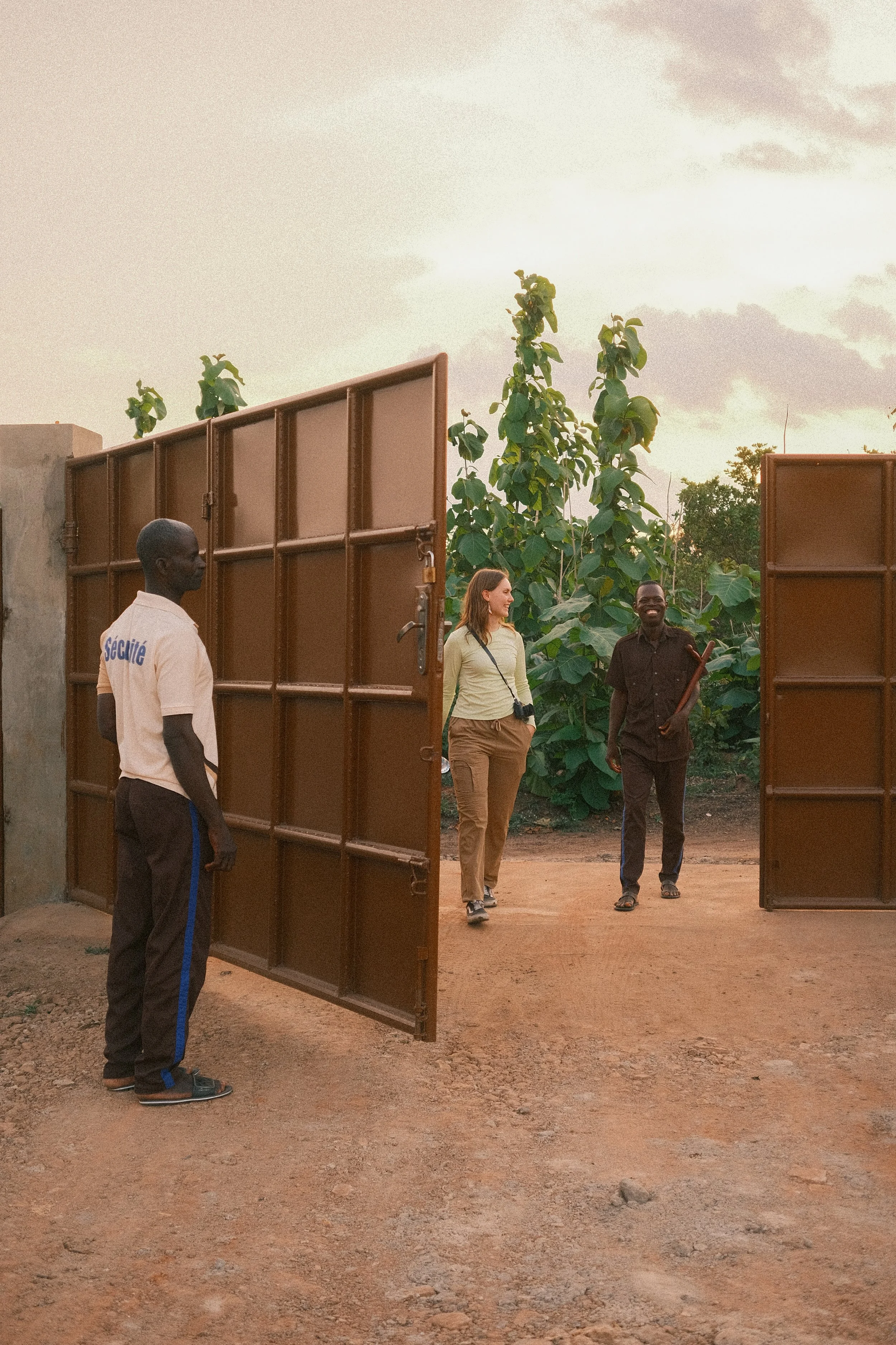 A woman and a man walk out of a large open brown gate on a dirt pathway, with green trees in the background during sunset. An attendant in a white uniform with blue accents stands near the gate.