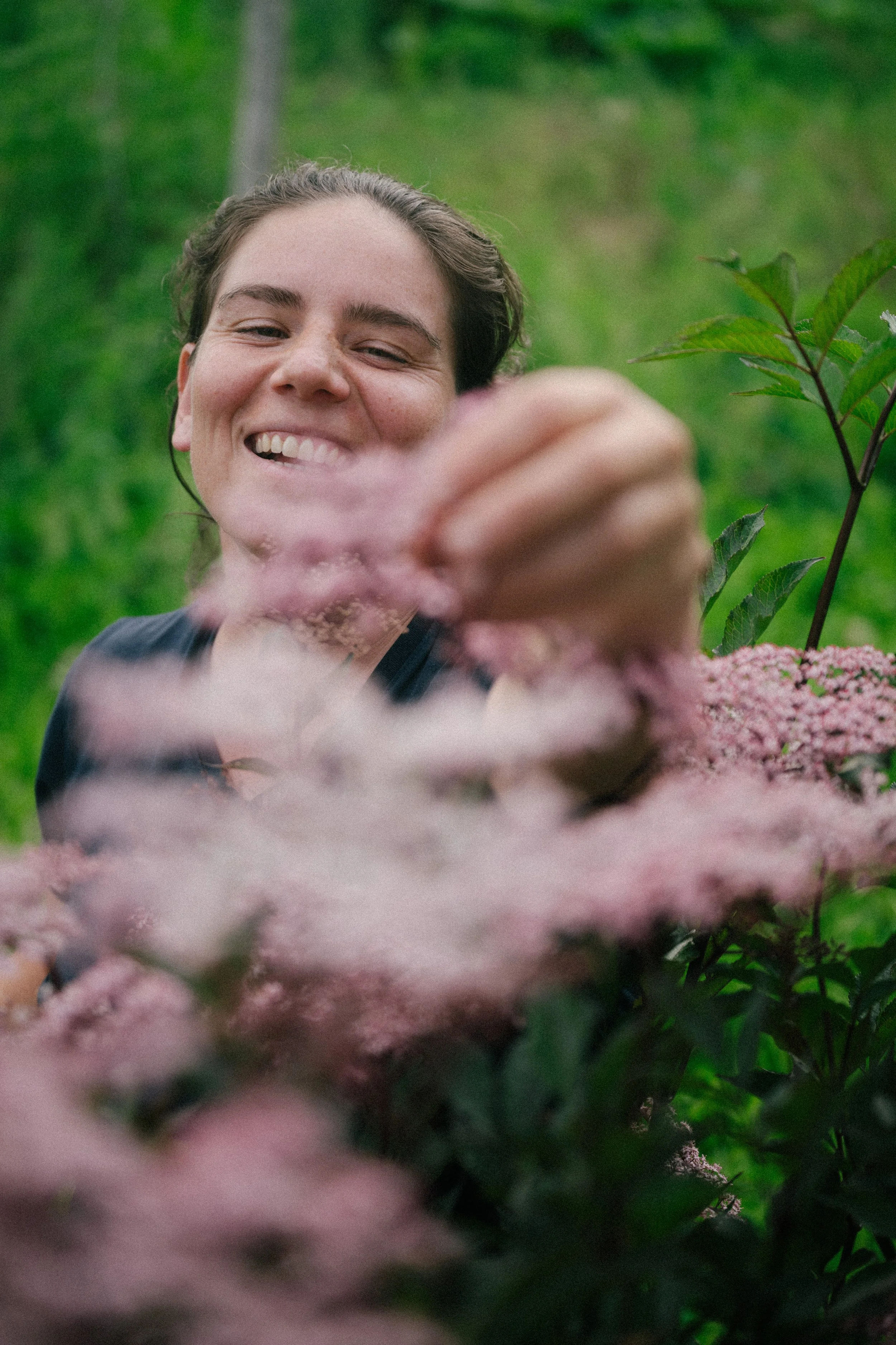 A smiling person outdoors surrounded by green foliage and pink flowers, reaching toward the camera.