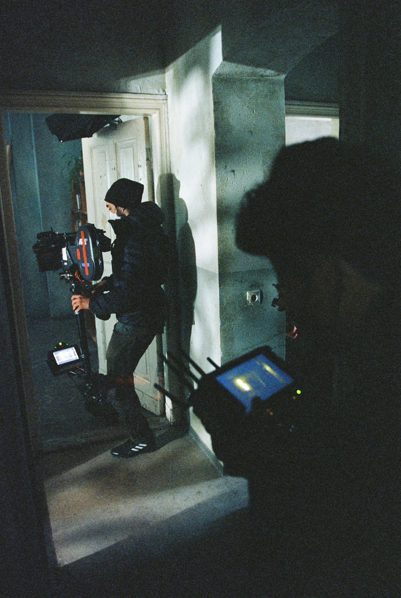 Filmmaker with camera and monitor inside a dimly lit building, with another person partially visible.