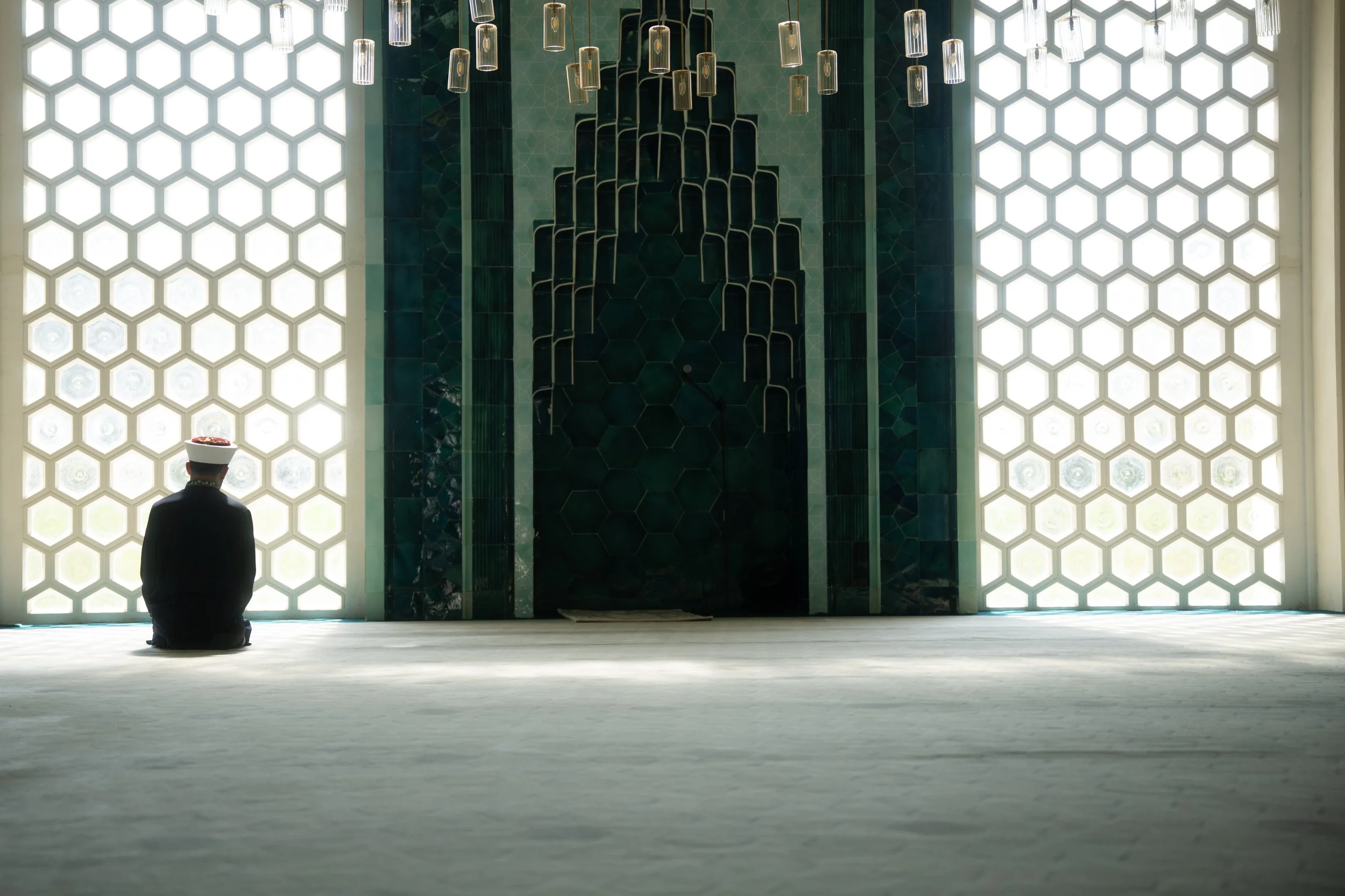 A person sitting in prayer in a mosque, facing a large green and black tiled mihrab with hexagonal windows on both sides, and hanging lights above.