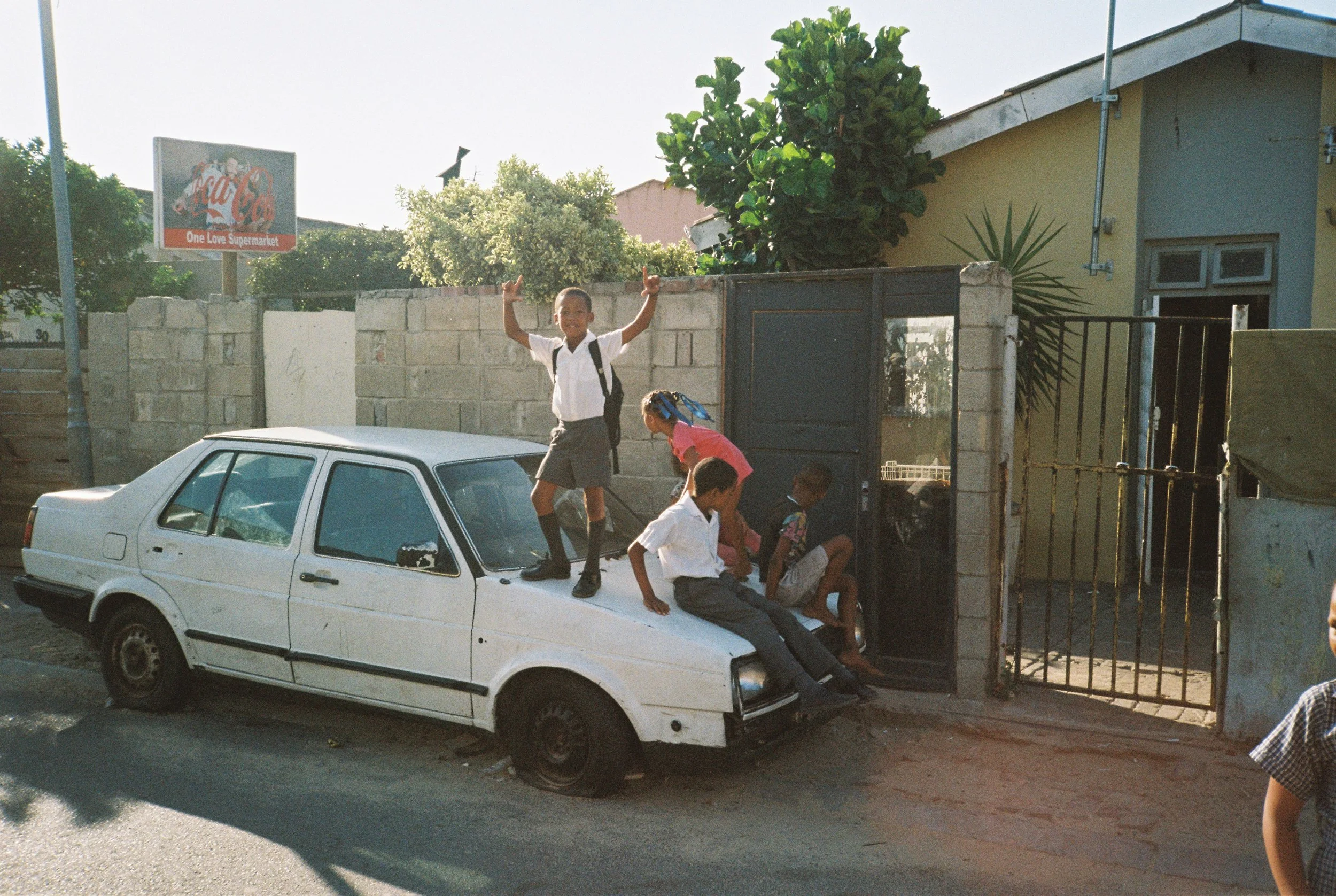 A group of children playing on and around a white car parked on the street, with some sitting on the car and one boy standing on top with arms raised, in front of a gate and wall with trees and a building in the background.