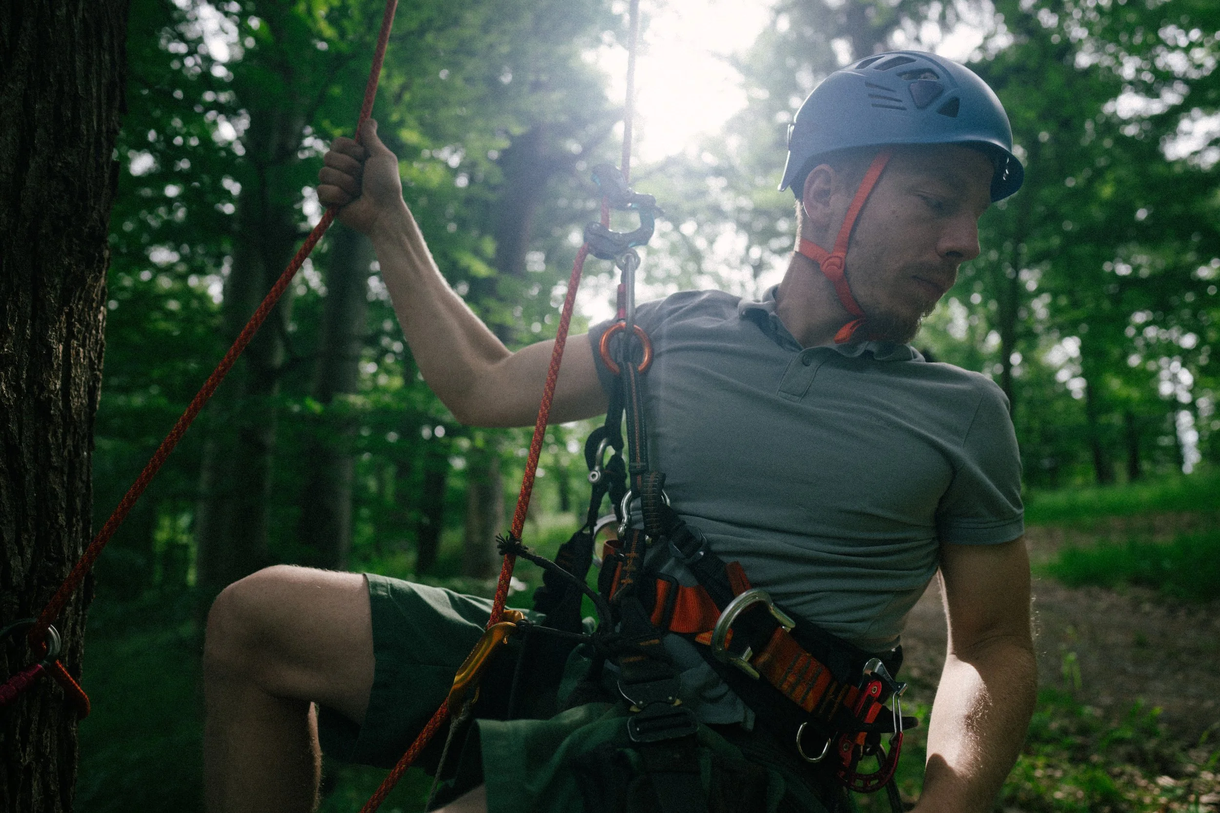 A man wearing a climbing helmet and safety gear, sitting in a forest, holding a climbing rope.