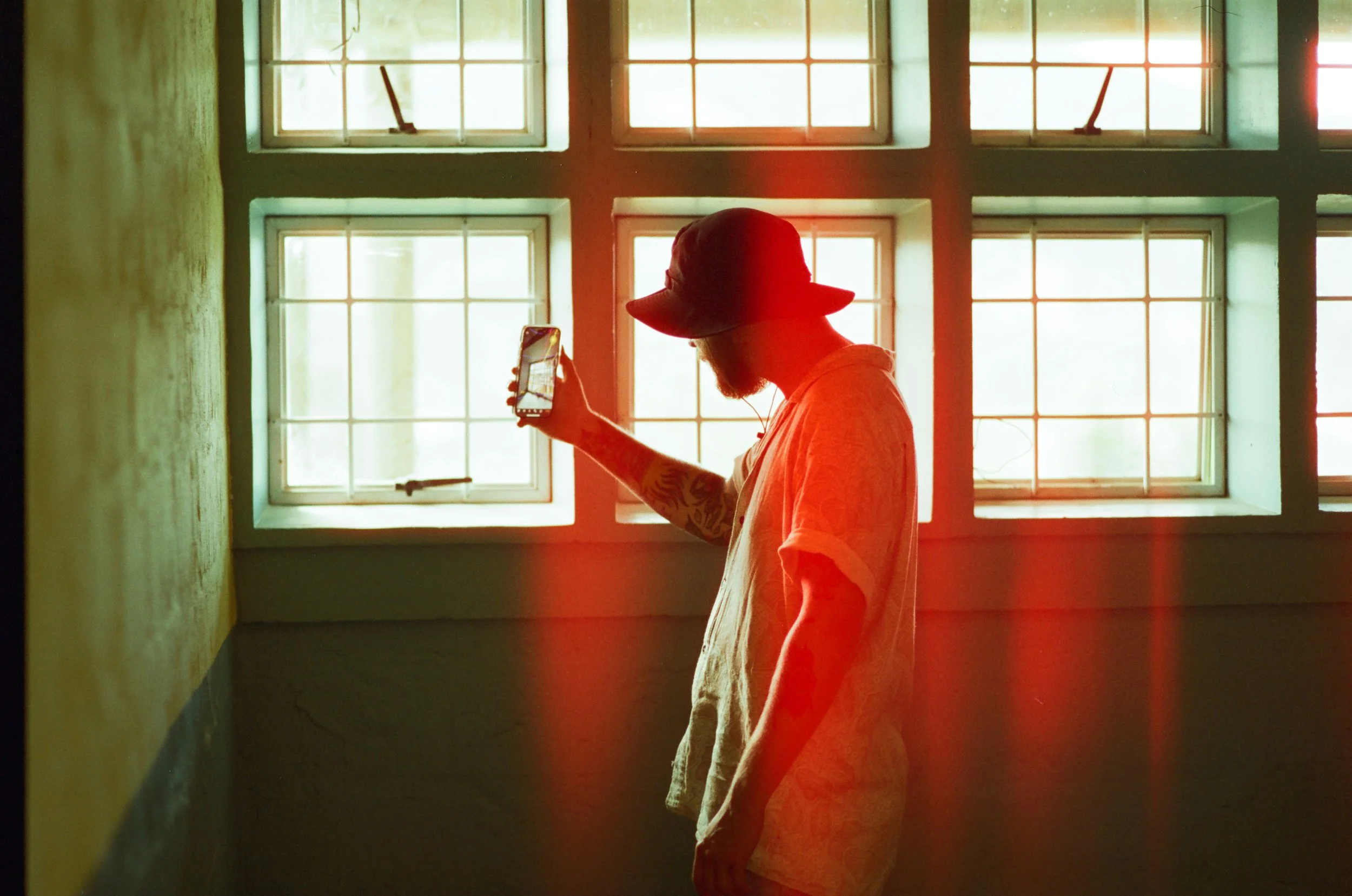 A man wearing a patterned shirt, hat, and glasses is standing indoors near a window, holding a smartphone with a red light glare across the image.