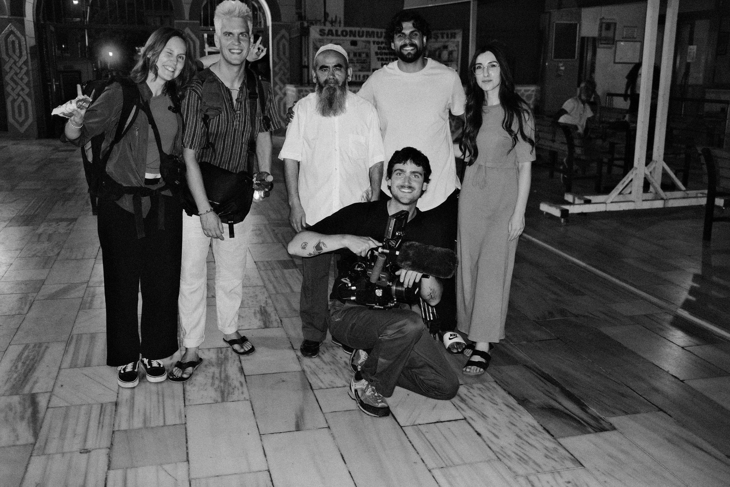 Group of seven people posing together indoors, including a man with a camera kneeling in front. Four women and two men stand behind, with different expressions, in a casual setting.