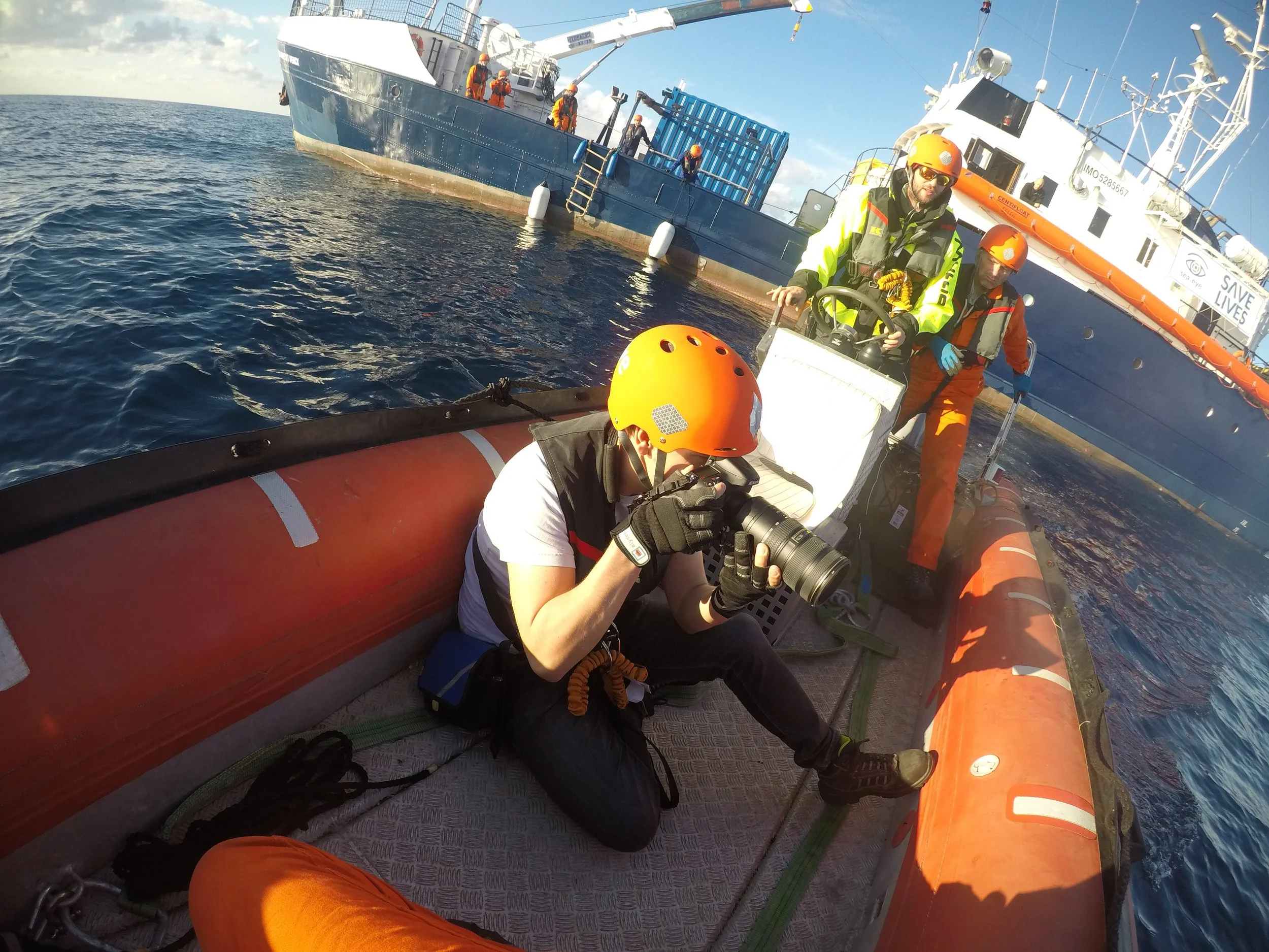 Team of rescue personnel on an orange inflatable boat taking photos during a maritime rescue operation, with large ships in the background on the water.
