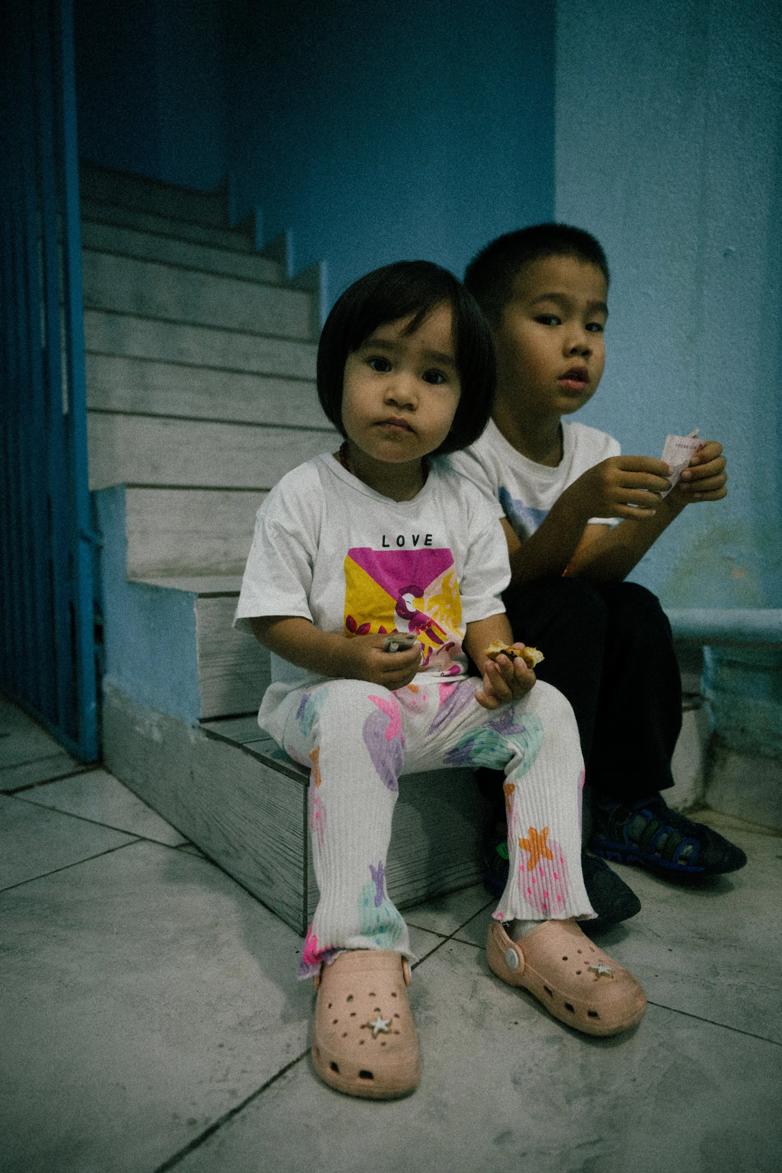Two young children sitting on stairs against a blue wall inside a home, with one girl in front holding snacks and an older boy behind holding a card.
