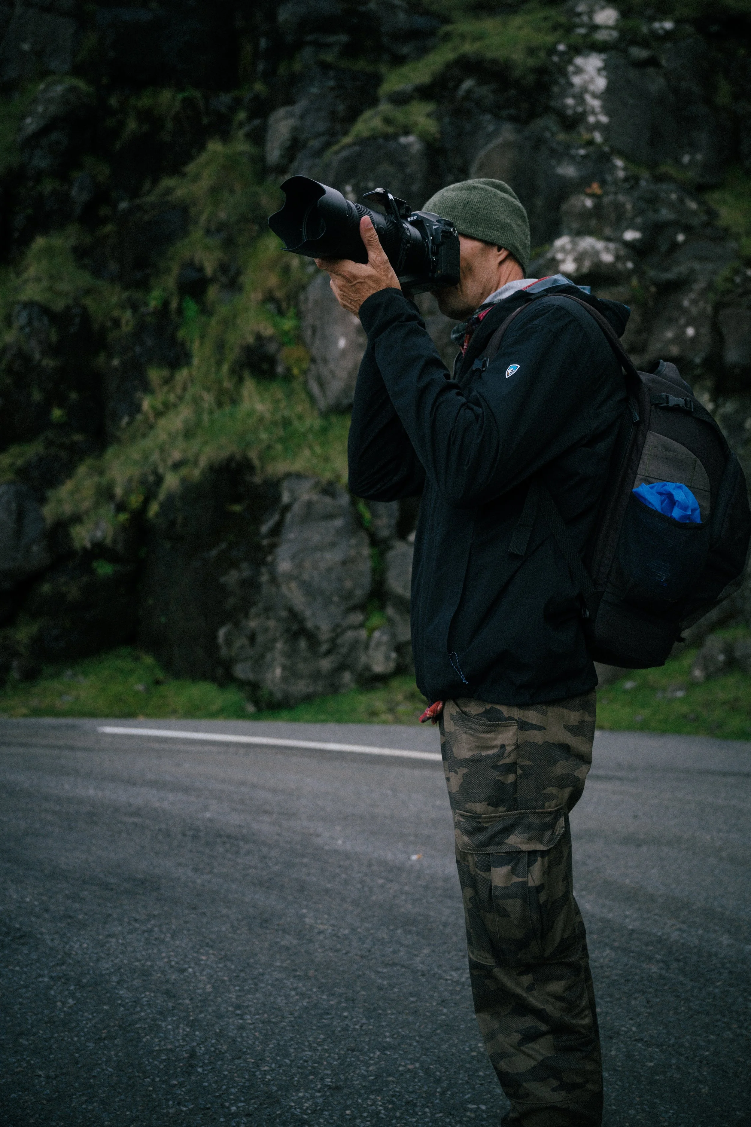A man wearing a green beanie and camouflage pants taking a photograph with a camera on a deserted road with rocky and grassy terrain in the background.