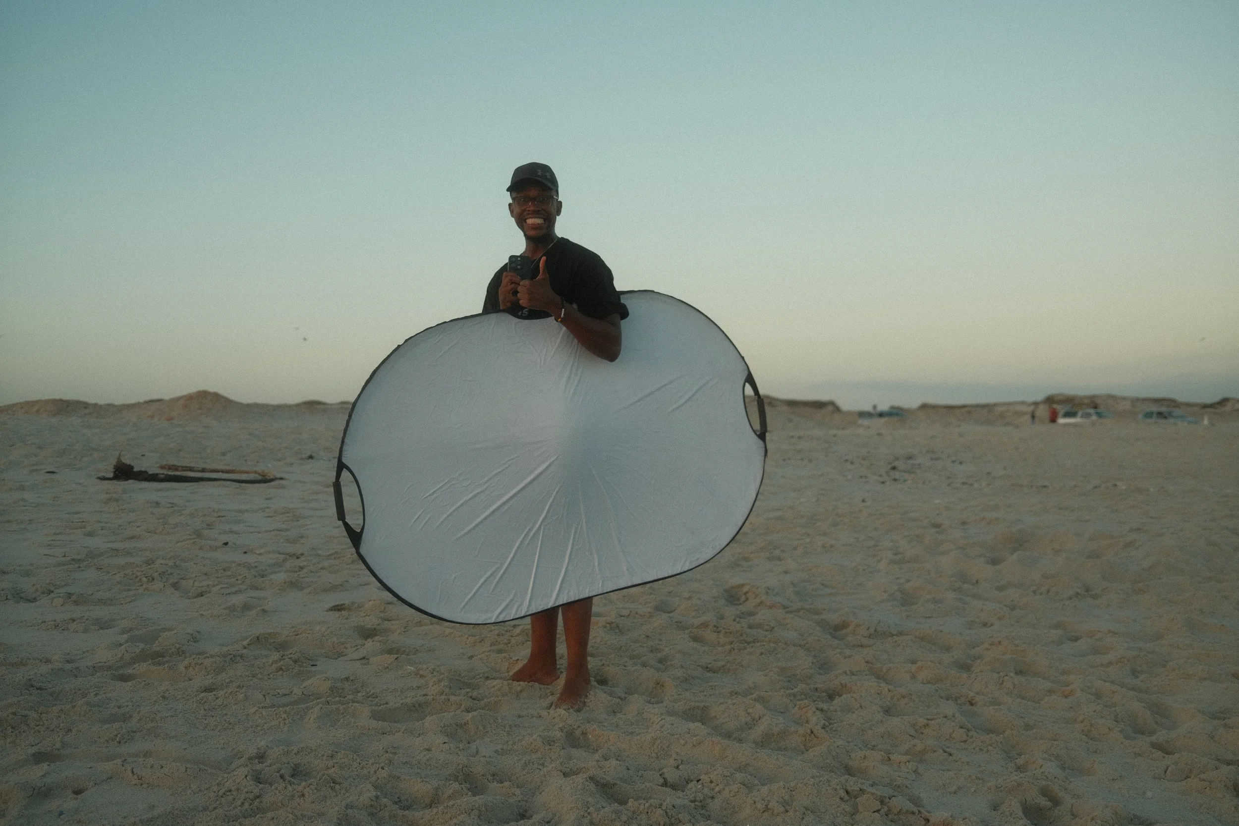 A person standing barefoot on a sandy beach holding a large photography reflector, smiling and giving a thumbs-up, with the sky and distant cars in the background.