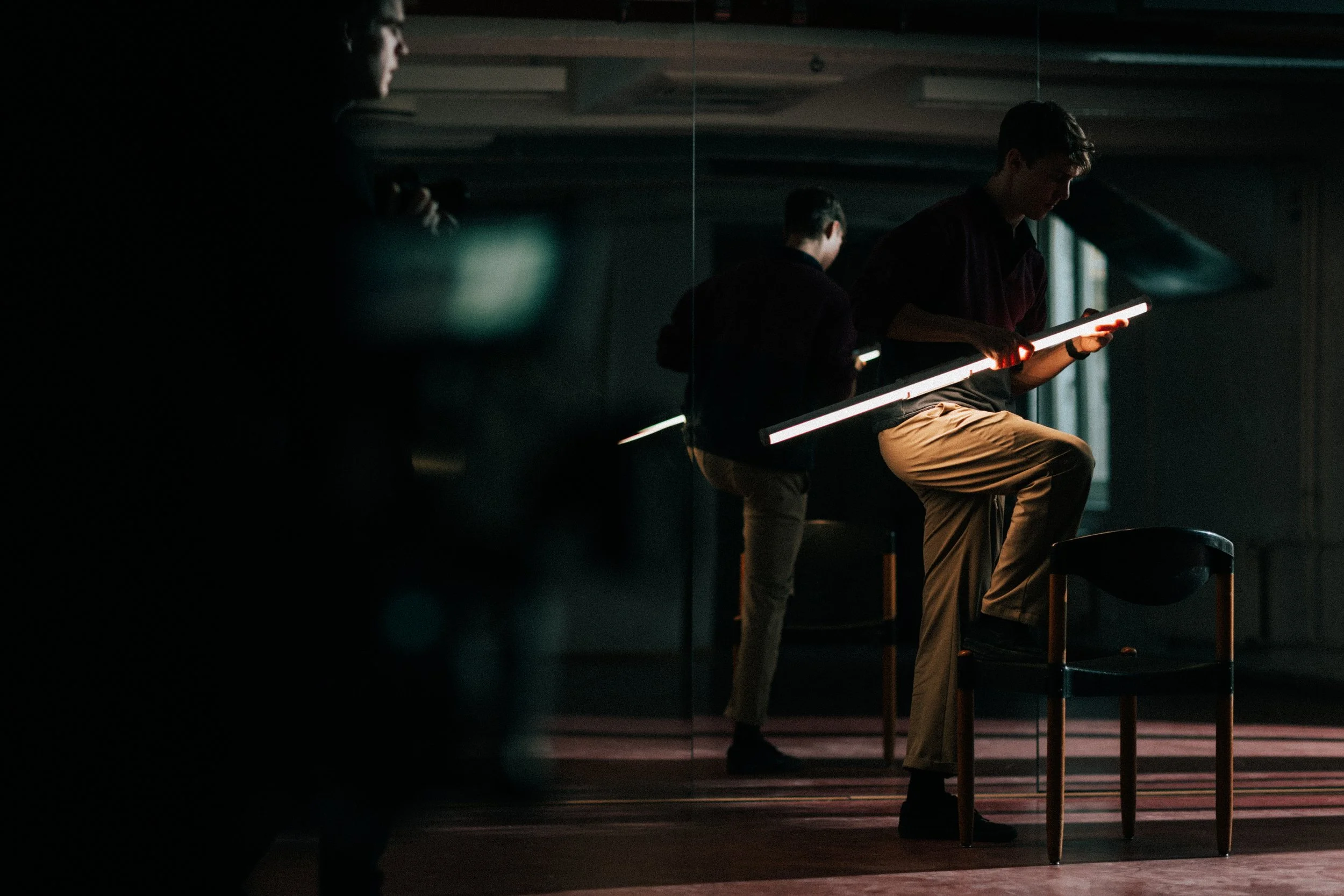 Two men practicing ballet in a dimly lit studio, each holding a lighted ballet bar while standing on chairs.